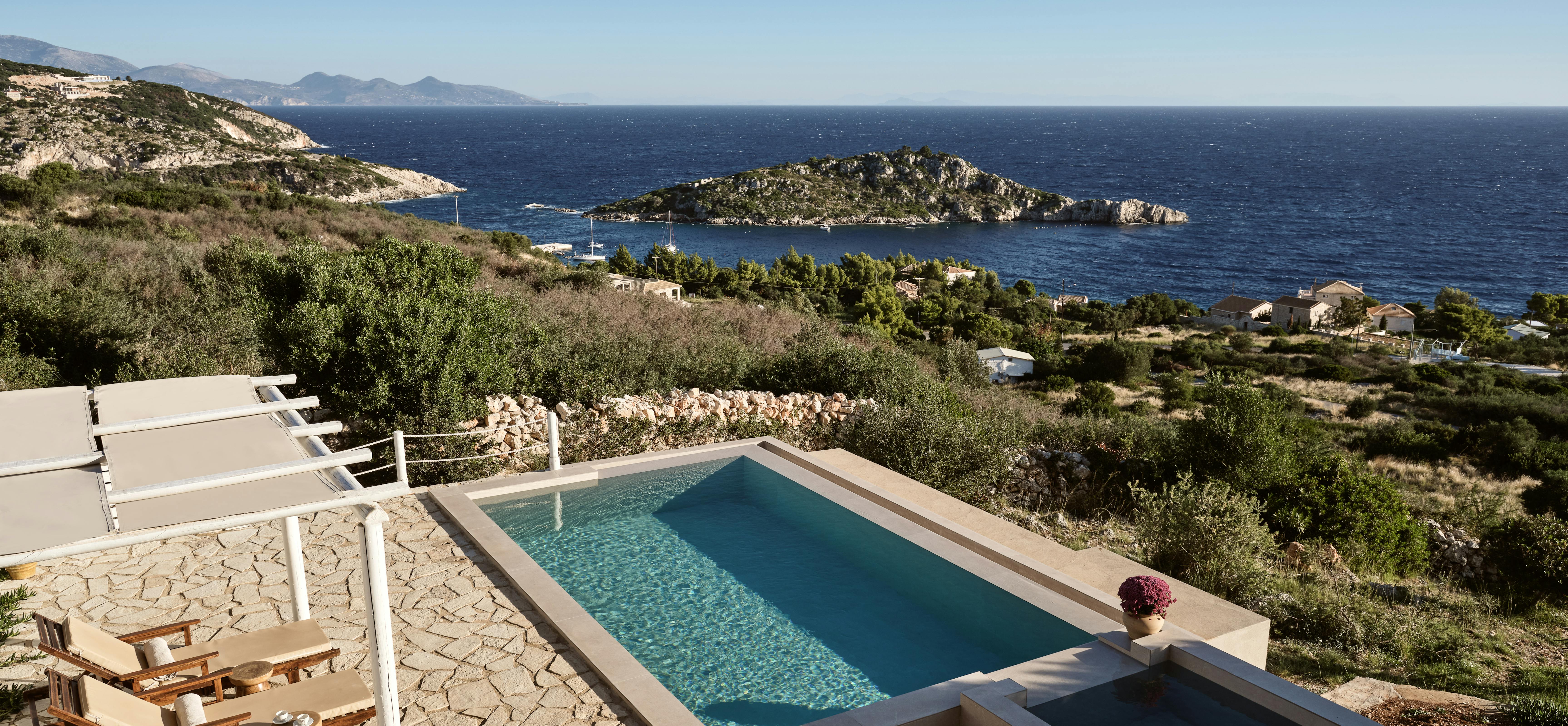 A multi-level infinity pool complex built into the hillside features stone terracing, white pergolas providing shade over wooden loungers, and sweeping views across a protected bay with rocky islands to the mountainous coastline beyond.