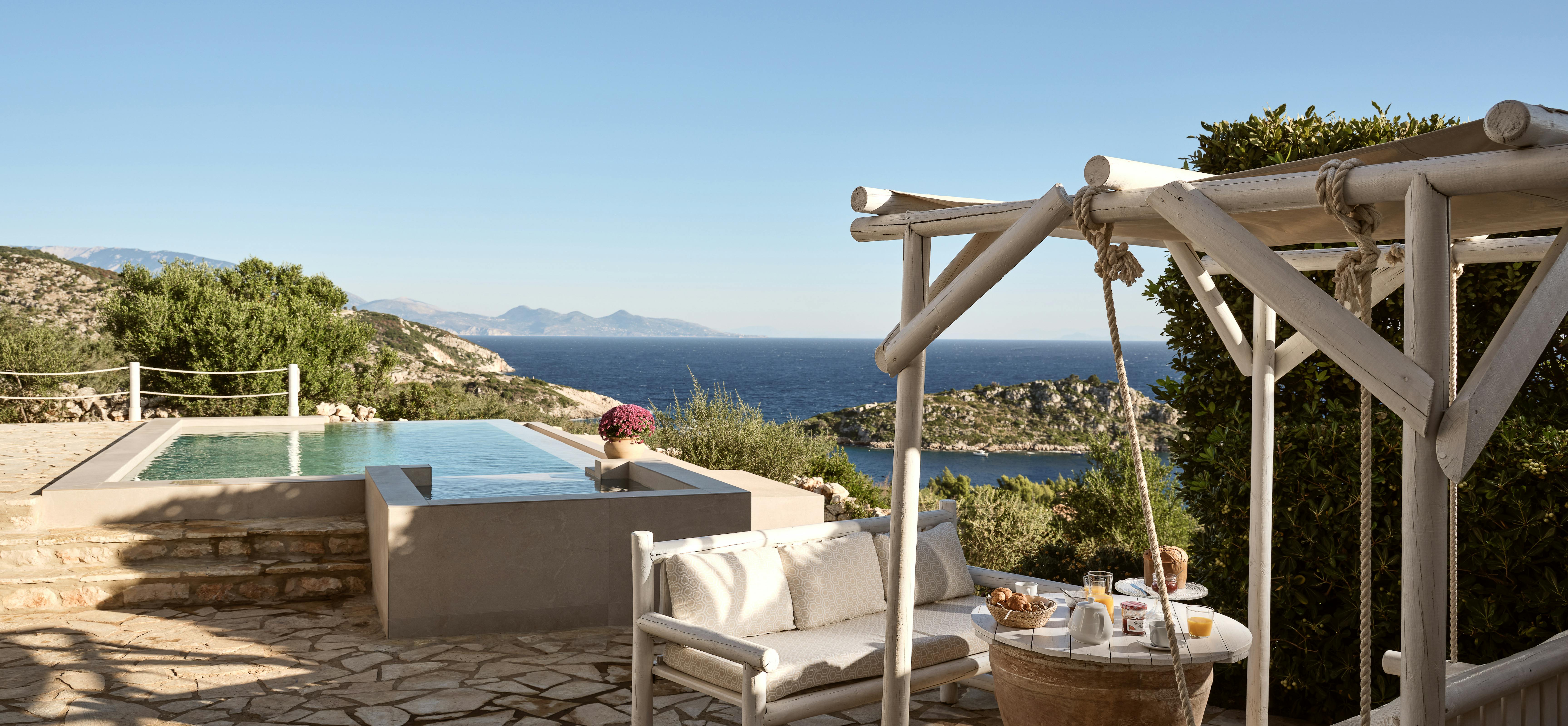 A white painted porch swing suspended from a rustic wooden pergola overlooks the multi-level infinity pool perched on a stone terrace, with the sparkling Mediterranean and rugged coastal hills creating a idyllic backdrop for outdoor relaxation.