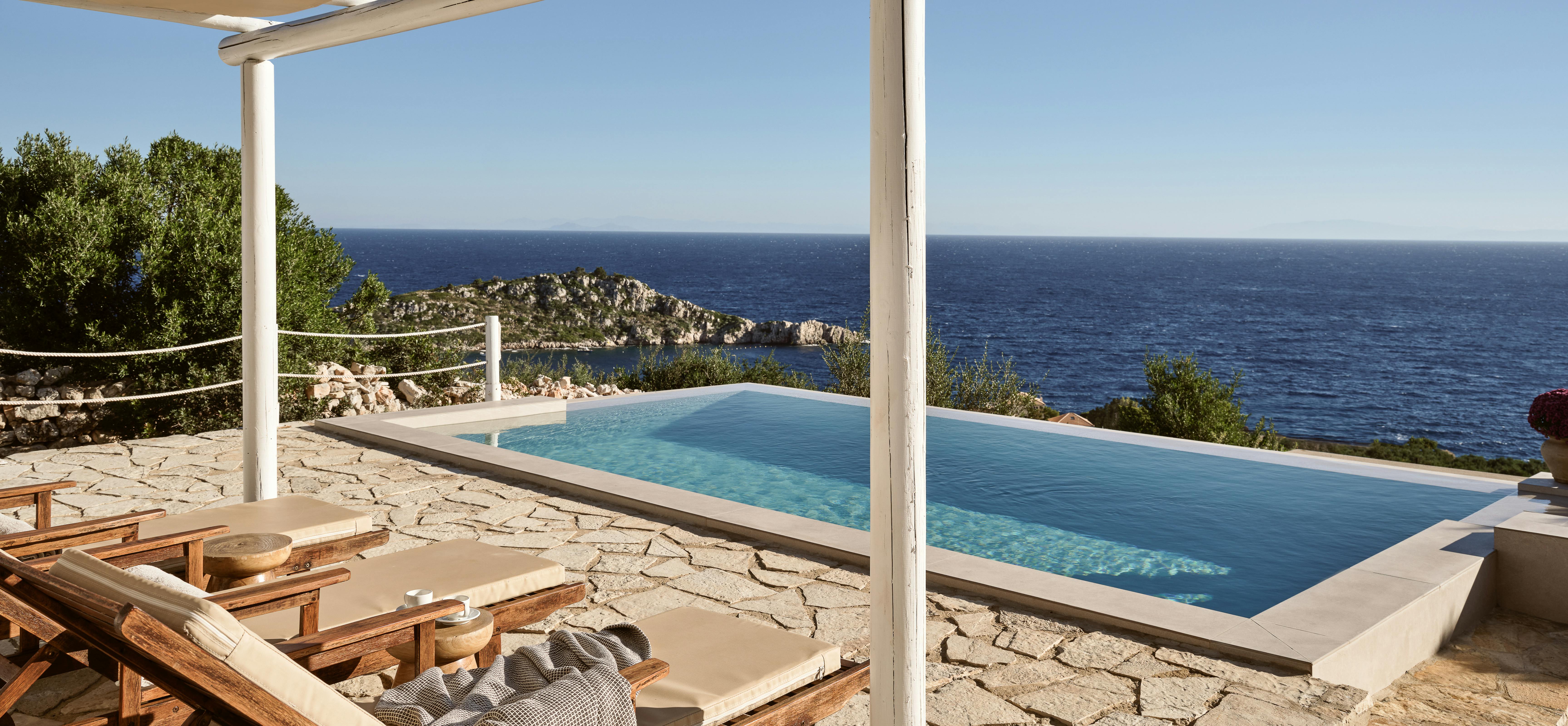 A white pergola with canvas shade panels shelters wooden sun loungers draped with textured throws on an irregular stone terrace, overlooking an infinity pool that appears to merge with the deep blue Mediterranean Sea and a dramatic rocky islet jutting from the water below.