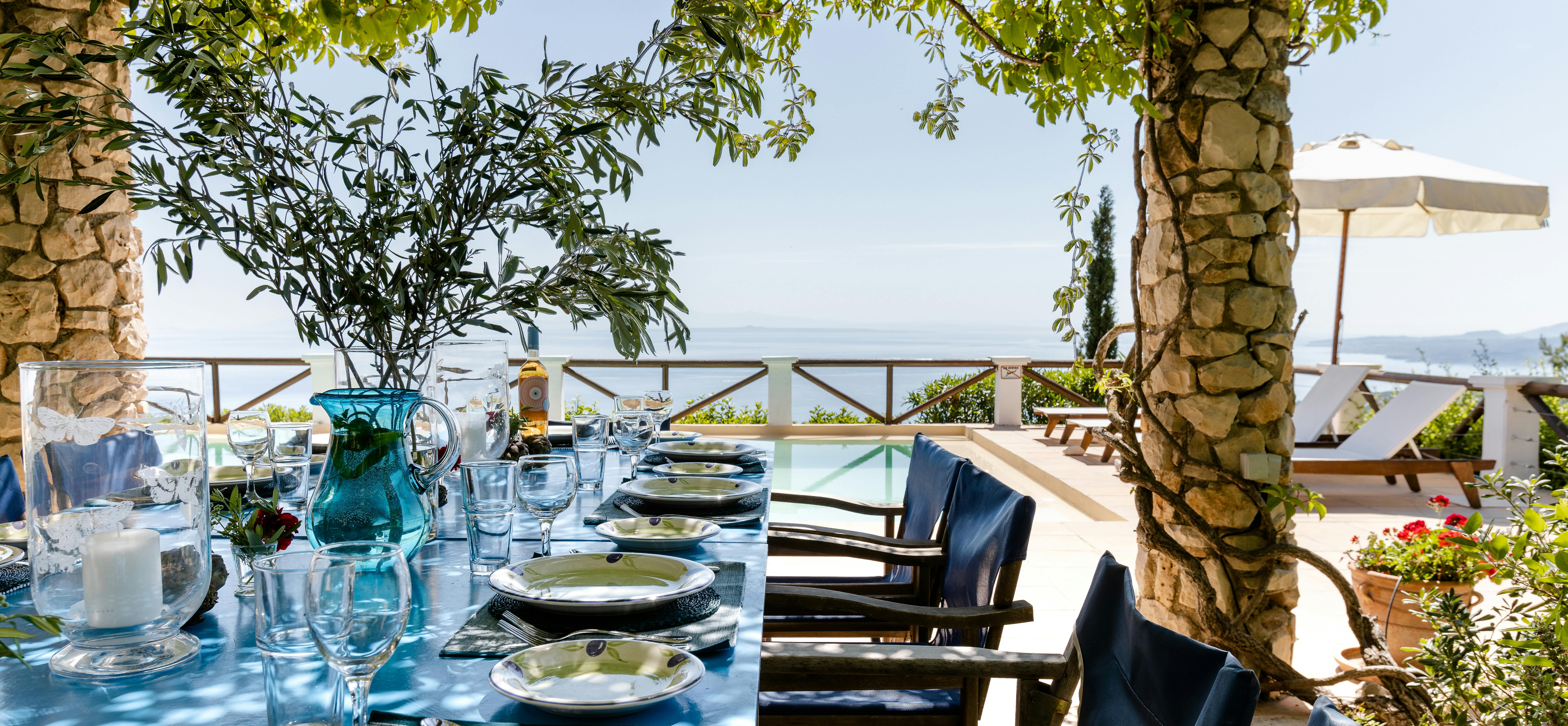 A vine-draped dining pergola frames an inviting outdoor table covered in bright blue cloth and set with olive-patterned dishes and crystal stemware, with blue director's chairs arranged along both sides overlooking an infinity pool and the shimmering sea beyond under dappled sunlight.
