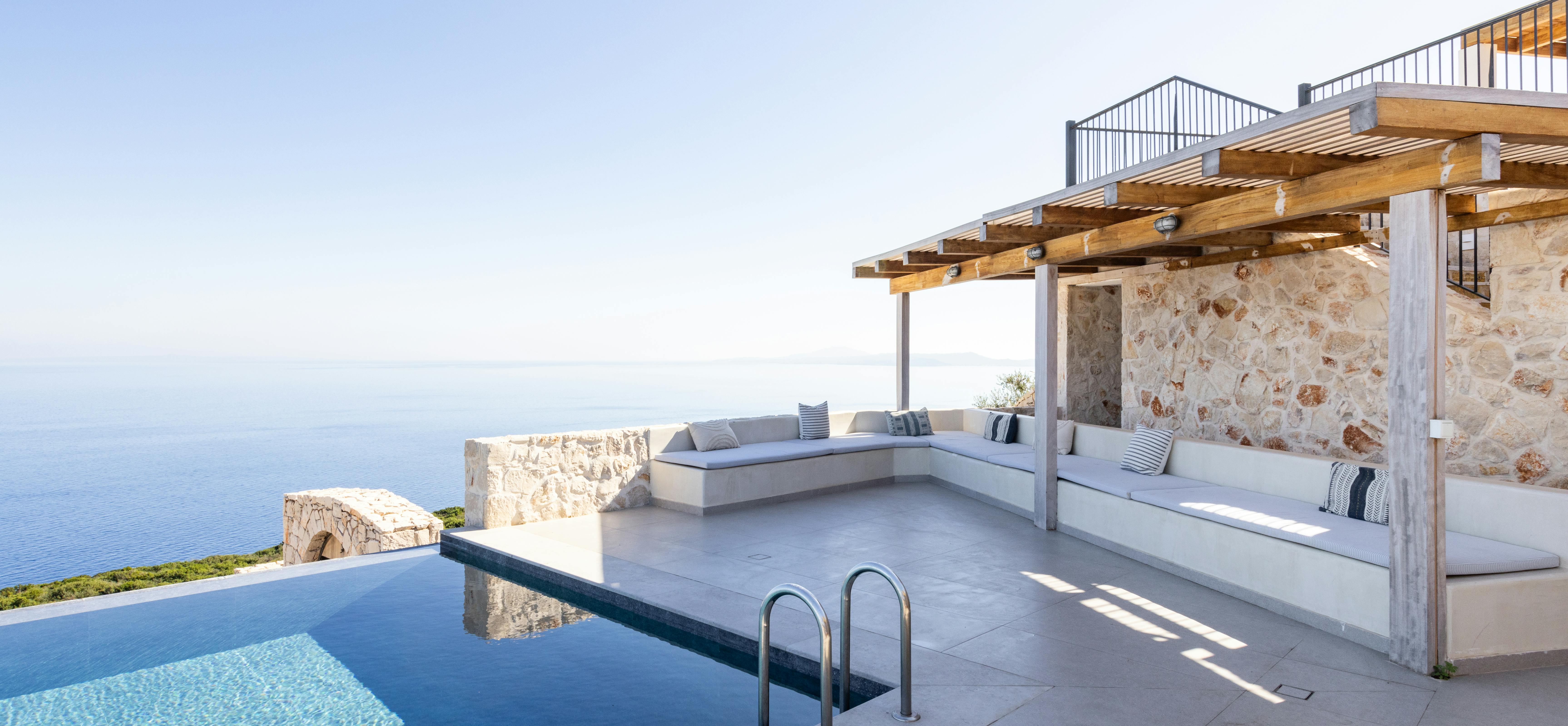 Stone villa pool terrace with built-in seating under pergola and infinity edge overlooking sea.