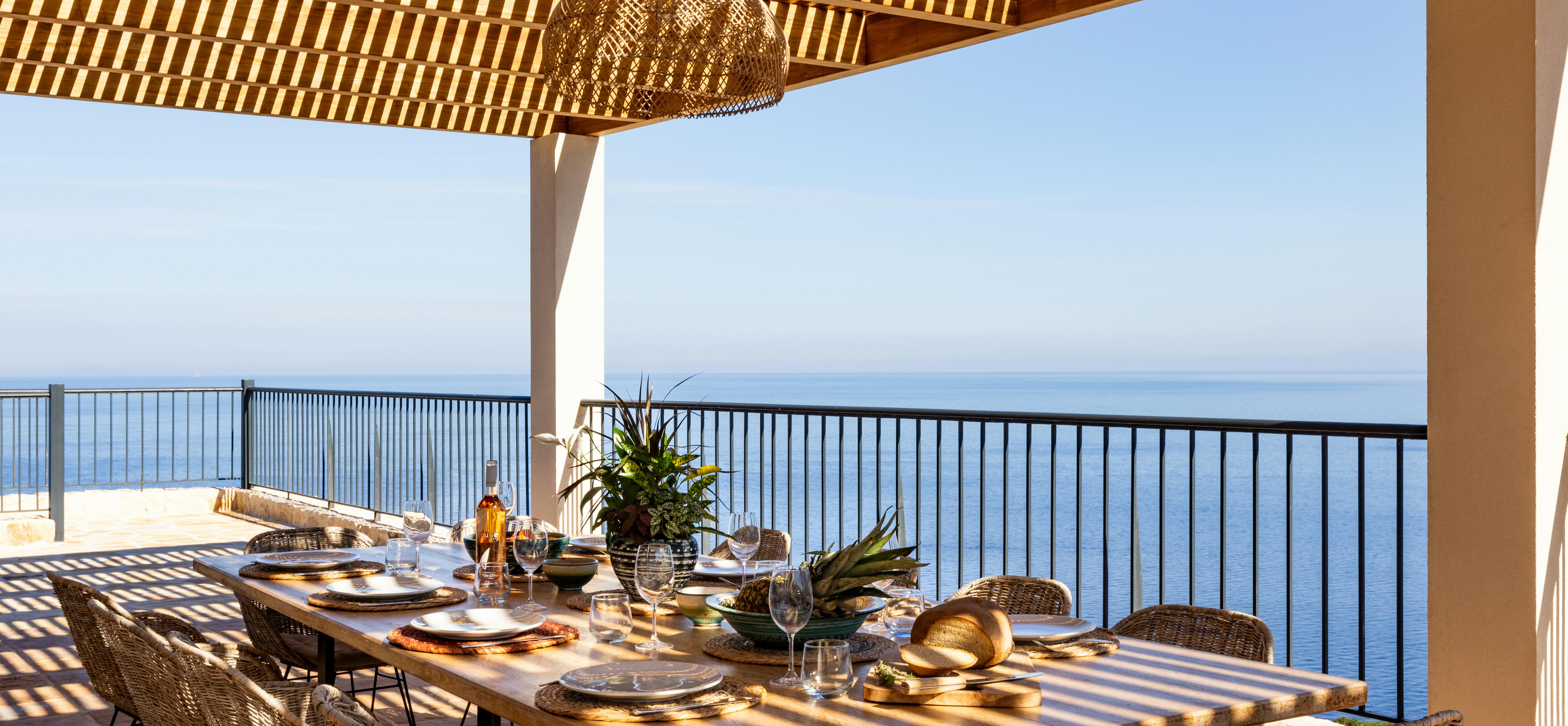 Outdoor dining terrace with wooden slatted pergola, woven chairs, and panoramic ocean views.