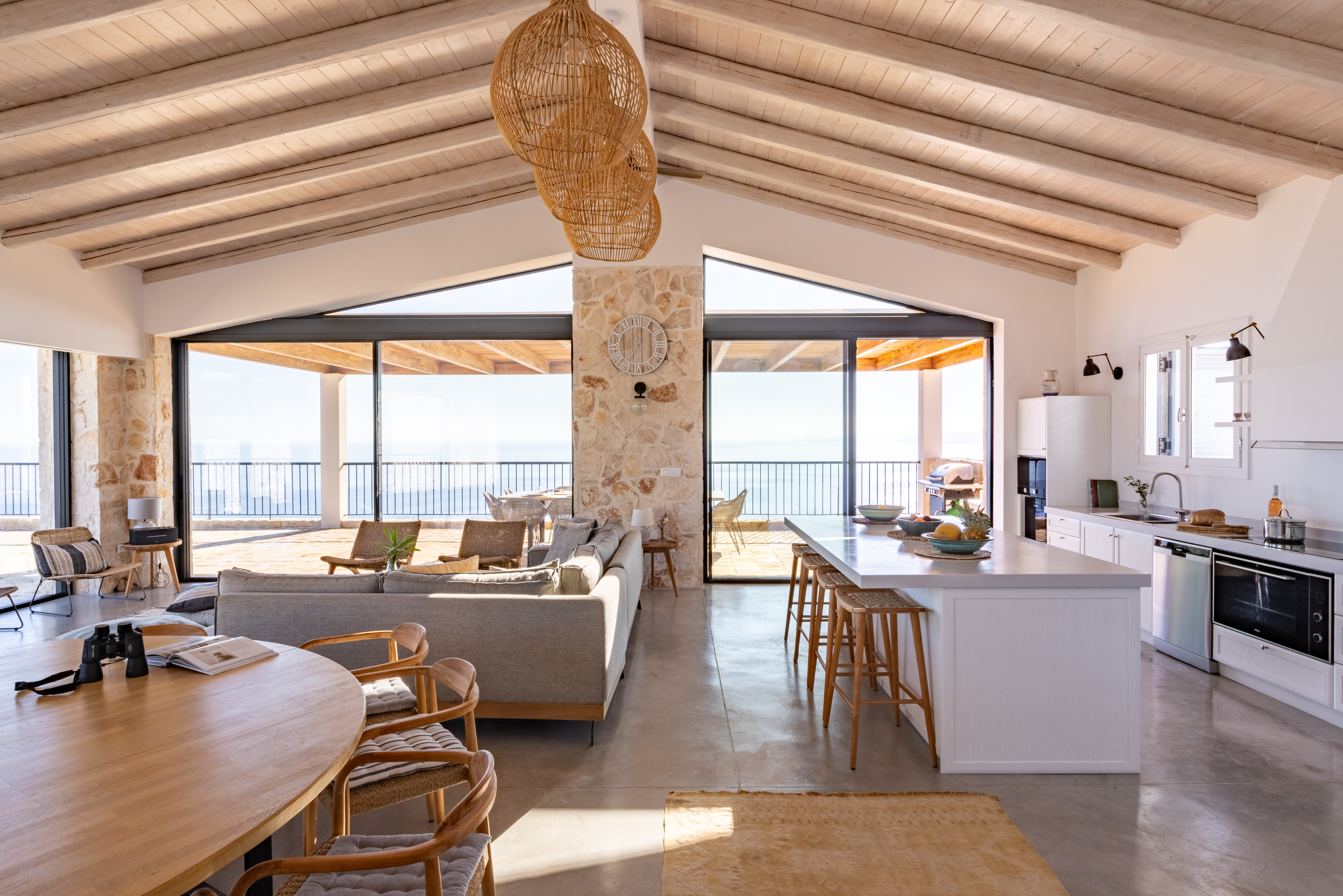 Open-plan living space with exposed wooden beams, stone pillar, kitchen island, and floor-to-ceiling windows overlooking the sea.
