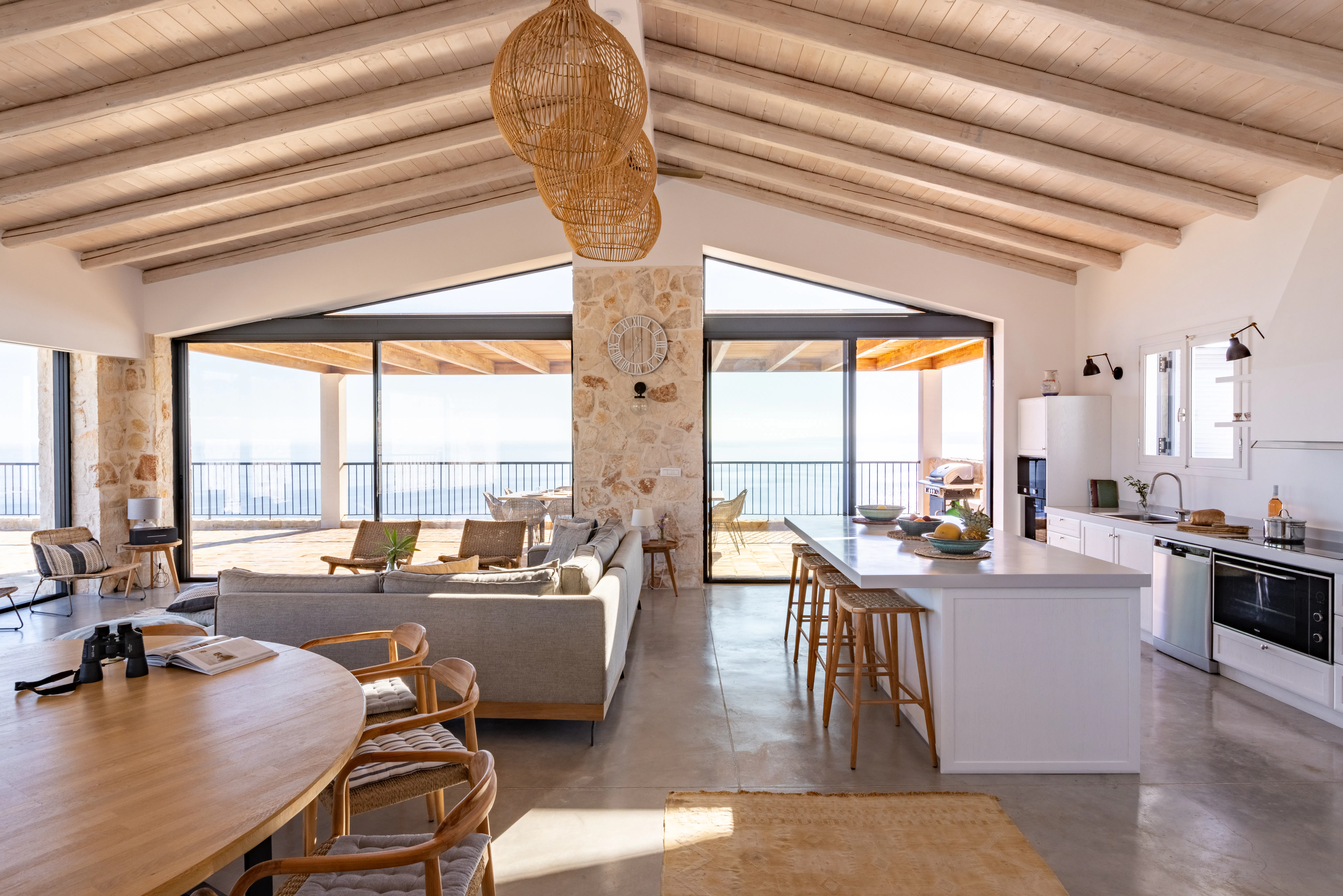 Open-plan living space with exposed wooden beams, stone pillar, kitchen island, and floor-to-ceiling windows overlooking the sea.