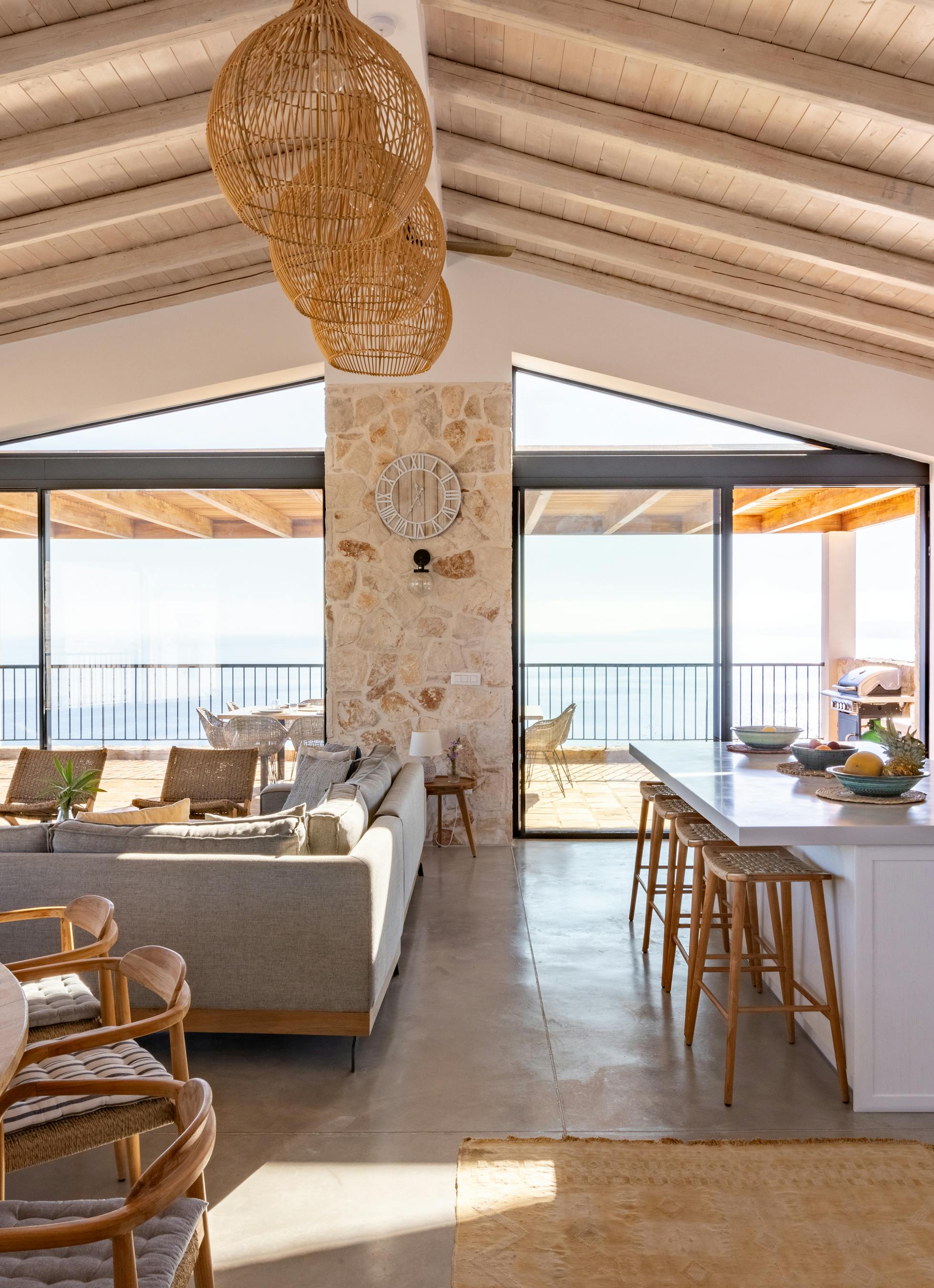Open-plan living space with exposed wooden beams, stone pillar, kitchen island, and floor-to-ceiling windows overlooking the sea.