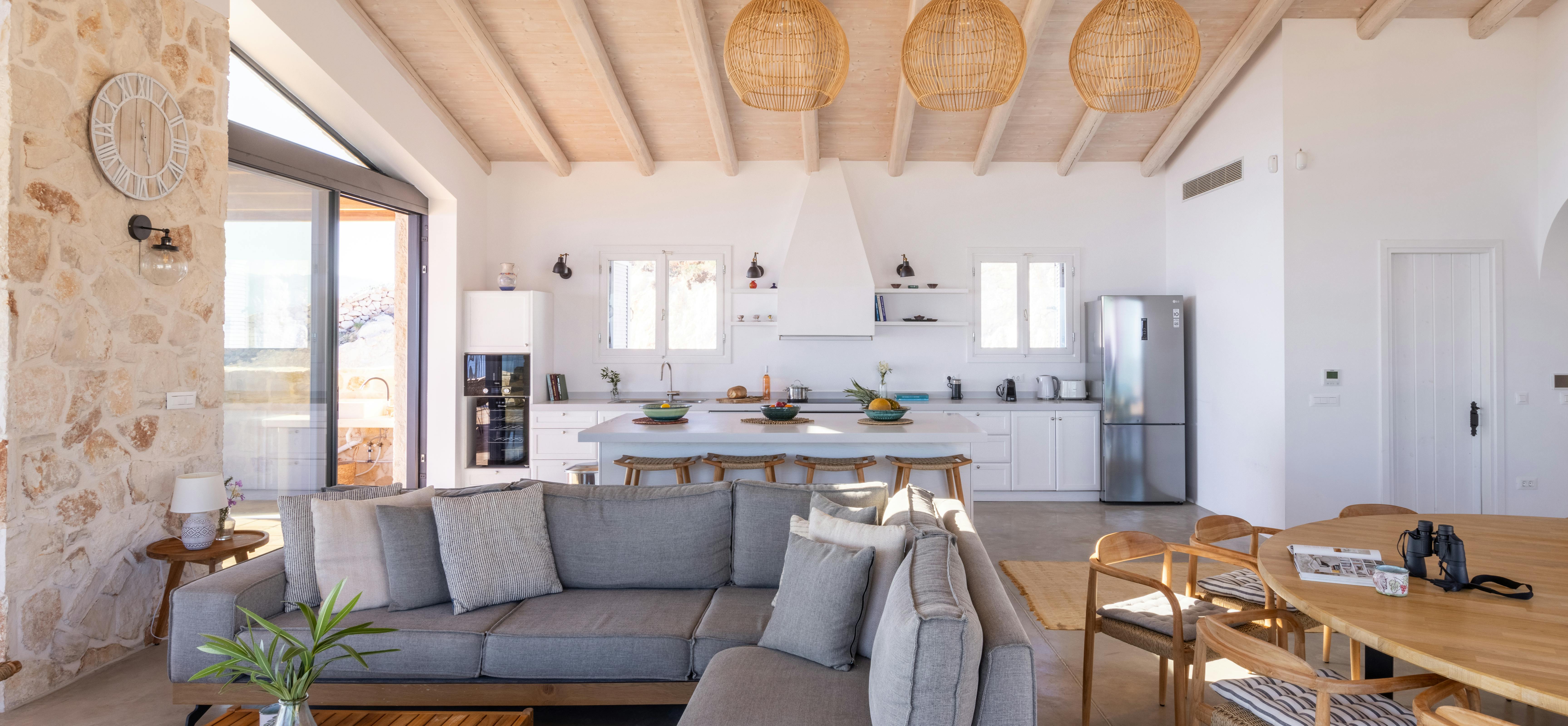 Bright open living area with grey sectional sofa, woven pendant lights, and white kitchen beyond stone accent wall.