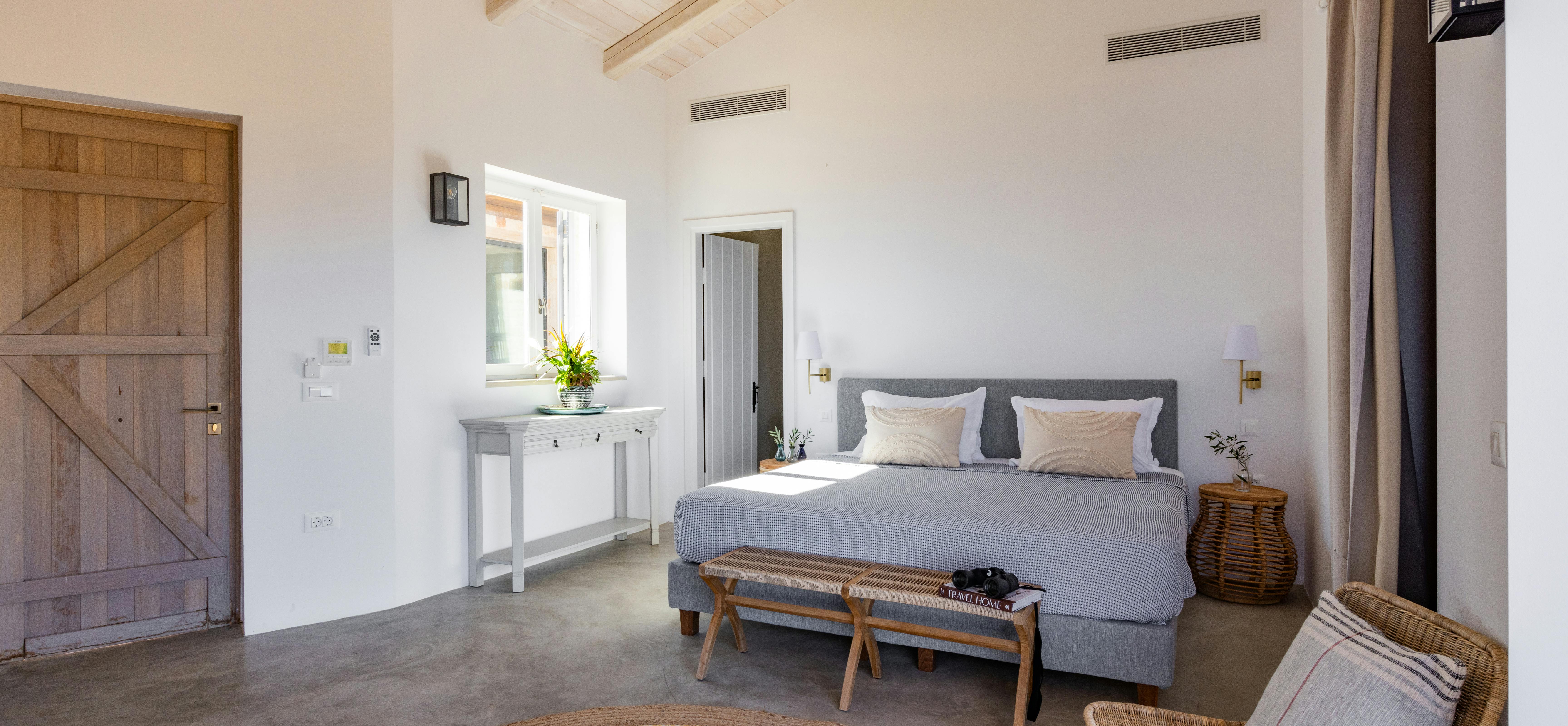 Spacious bedroom with vaulted wood ceiling, grey bed, rustic bench, and jute area rug.