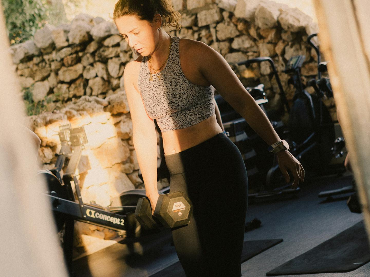 A woman in a grey patterned crop top and black leggings performs a lunge exercise with a dumbbell in a rustic gym with exposed stone walls and wooden beams. She's positioned on black exercise mats, with cardio equipment visible in the background. Natural light streams in, creating warm tones throughout the Mediterranean-style workout space.