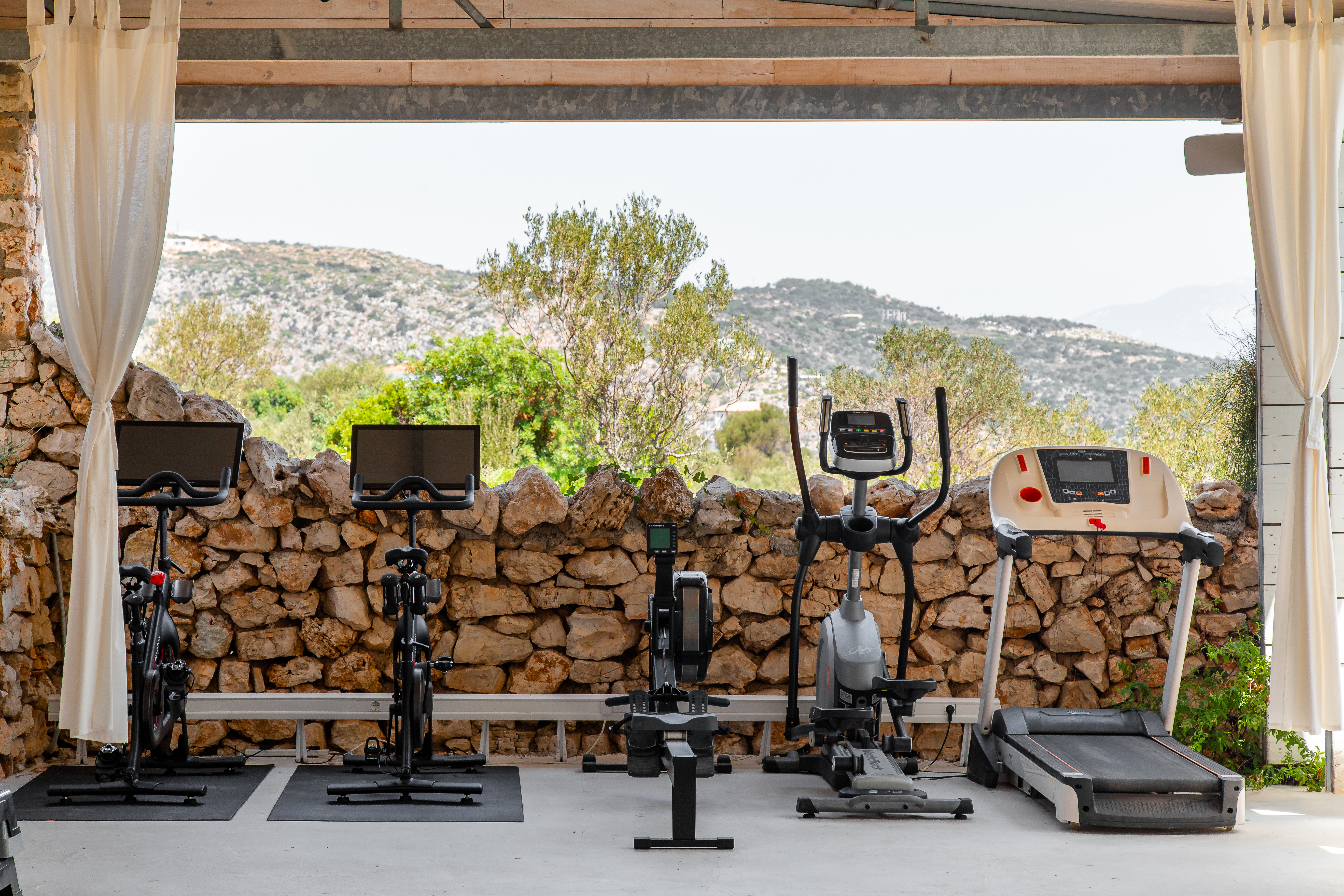 An outdoor fitness area featuring a row of exercise equipment including two spin bikes, a rowing machine, and a treadmill arranged on black mats under a covered pavilion. The equipment faces a rustic stone wall backdrop with Mediterranean hillside views and olive trees visible beyond. Sheer beige curtains frame the sides of the open-air gym space, creating an elegant yet natural workout environment.