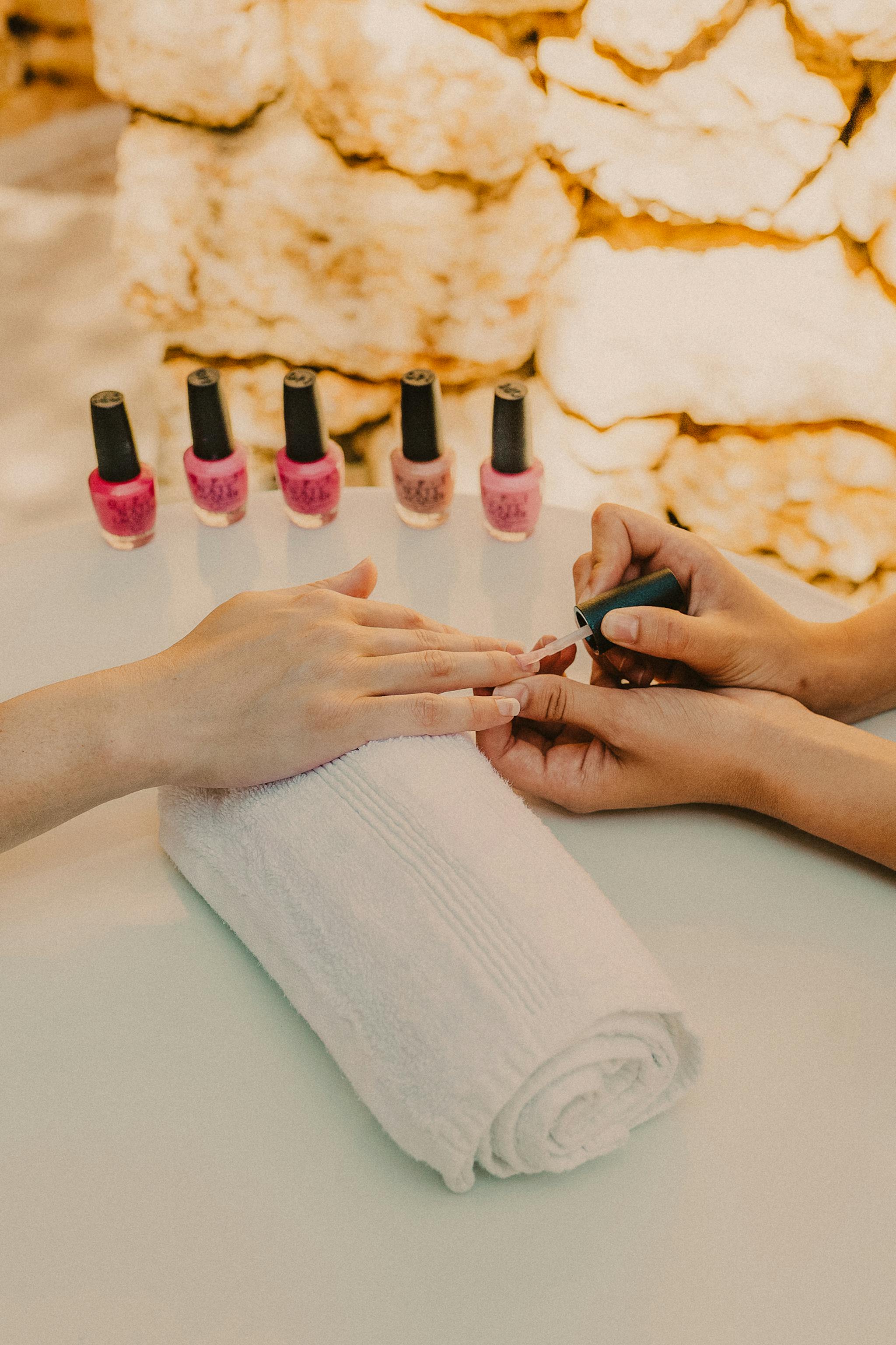 A manicurist applies nail polish to a client's hand during an outdoor manicure service. The client's hand rests on a white rolled towel on a light blue manicure table. Five bottles of pink and mauve nail polish are arranged in the background. The setting features a rustic stone wall backdrop, creating a serene spa atmosphere with natural lighting.