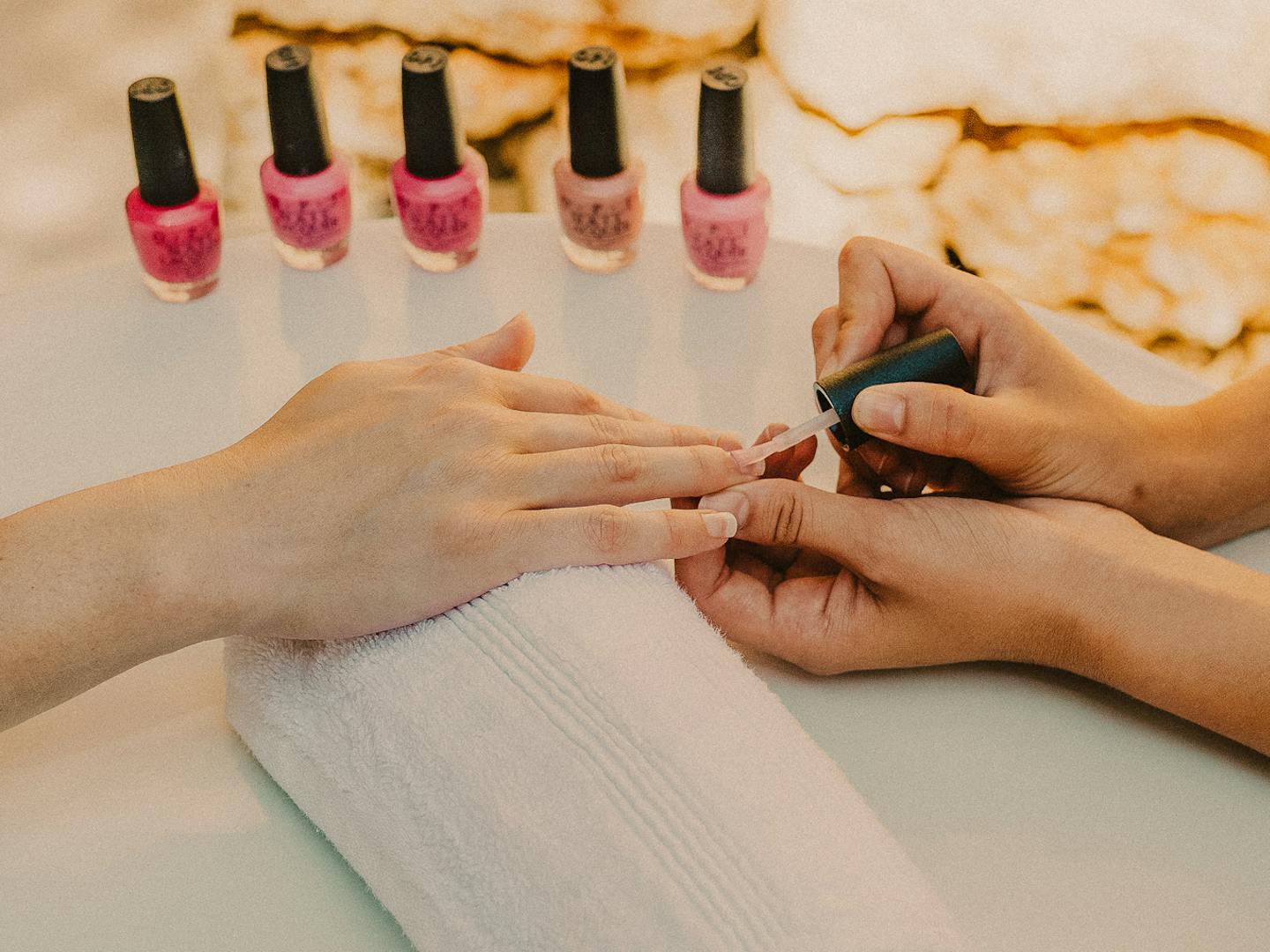 A manicurist applies nail polish to a client's hand during an outdoor manicure service. The client's hand rests on a white rolled towel on a light blue manicure table. Five bottles of pink and mauve nail polish are arranged in the background. The setting features a rustic stone wall backdrop, creating a serene spa atmosphere with natural lighting.