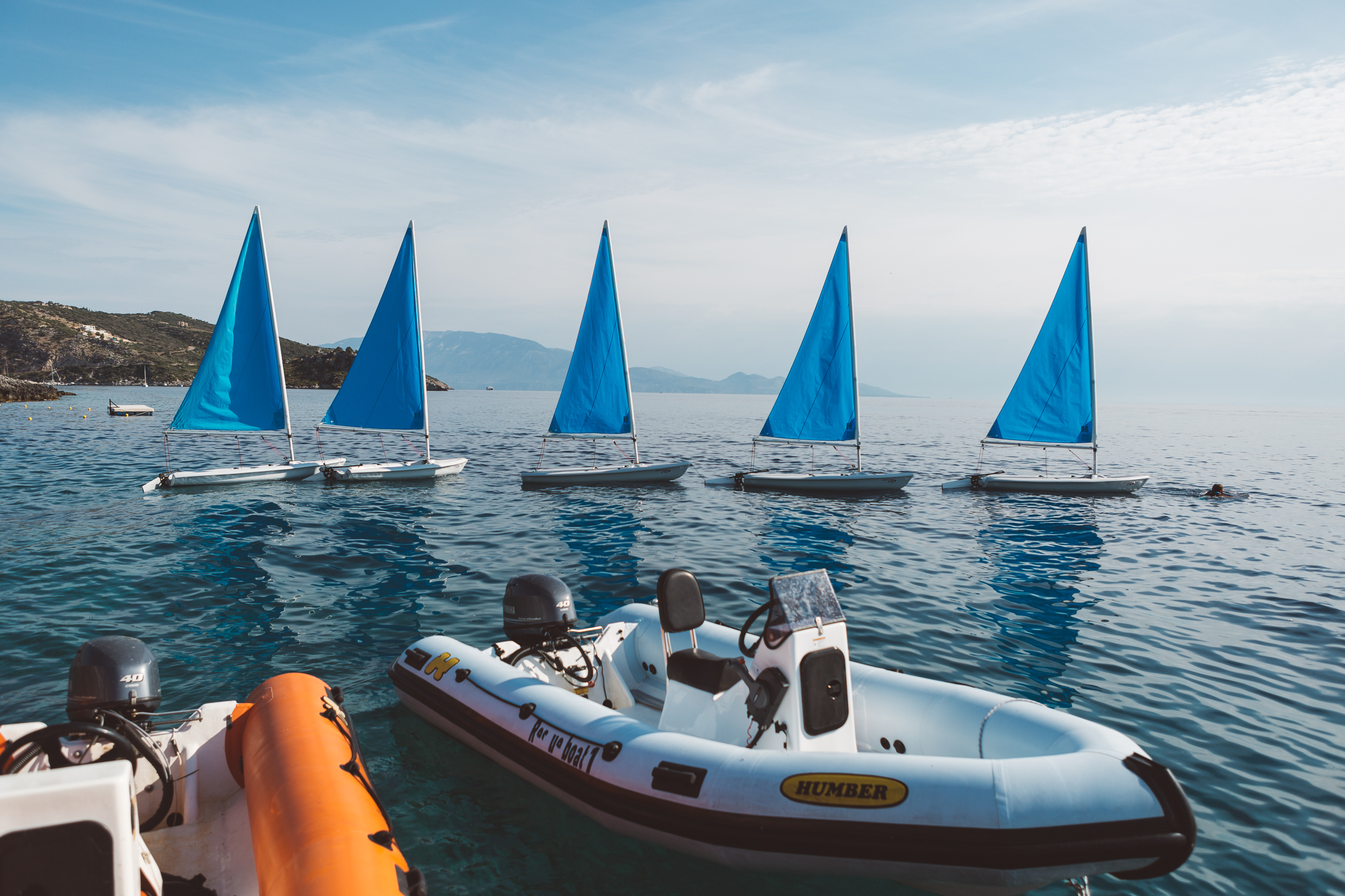 Five small sailboats with bright blue triangular sails lined up in a row on calm, deep blue water. In the foreground are inflatable dinghies with outboard motors, including a Humber brand boat. Mountains are visible in the hazy background under a partly cloudy sky, with a rocky coastline to the left.
