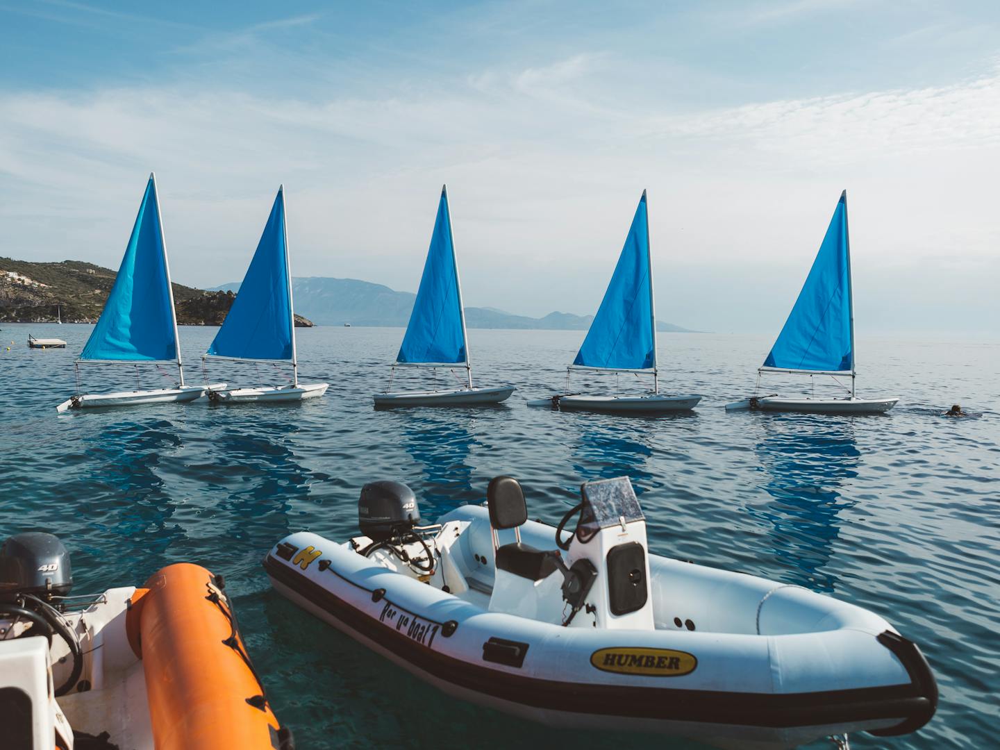 Five small sailboats with bright blue triangular sails lined up in a row on calm, deep blue water. In the foreground are inflatable dinghies with outboard motors, including a Humber brand boat. Mountains are visible in the hazy background under a partly cloudy sky, with a rocky coastline to the left.