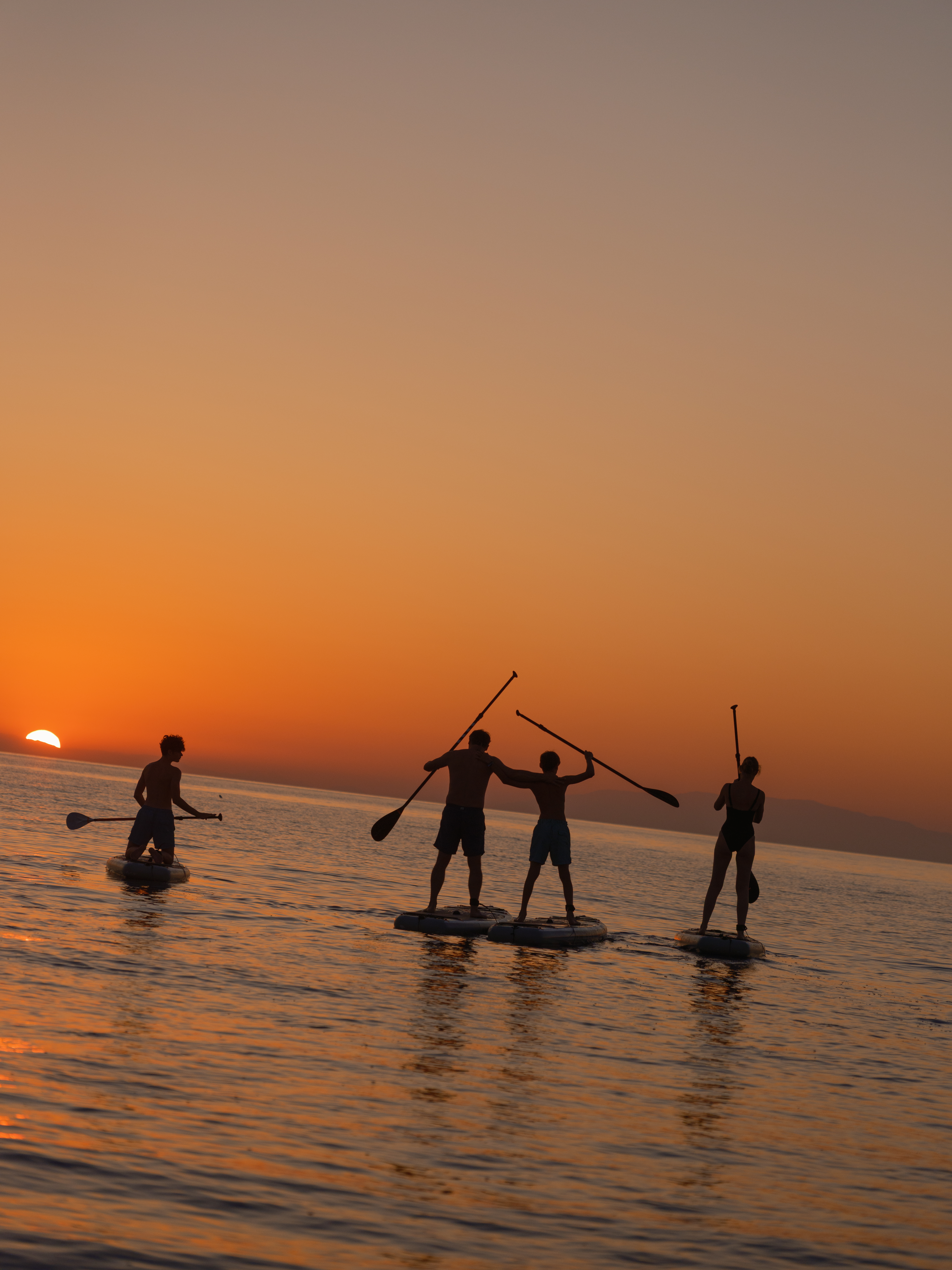 Silhouettes of four people stand-up paddleboarding at sunset on calm water. Three people are on paddleboards while one person stands in the shallow water. They're holding their paddles in various positions against a vibrant orange and amber sky as the sun sets on the horizon, creating dramatic backlighting and reflections on the water's surface.