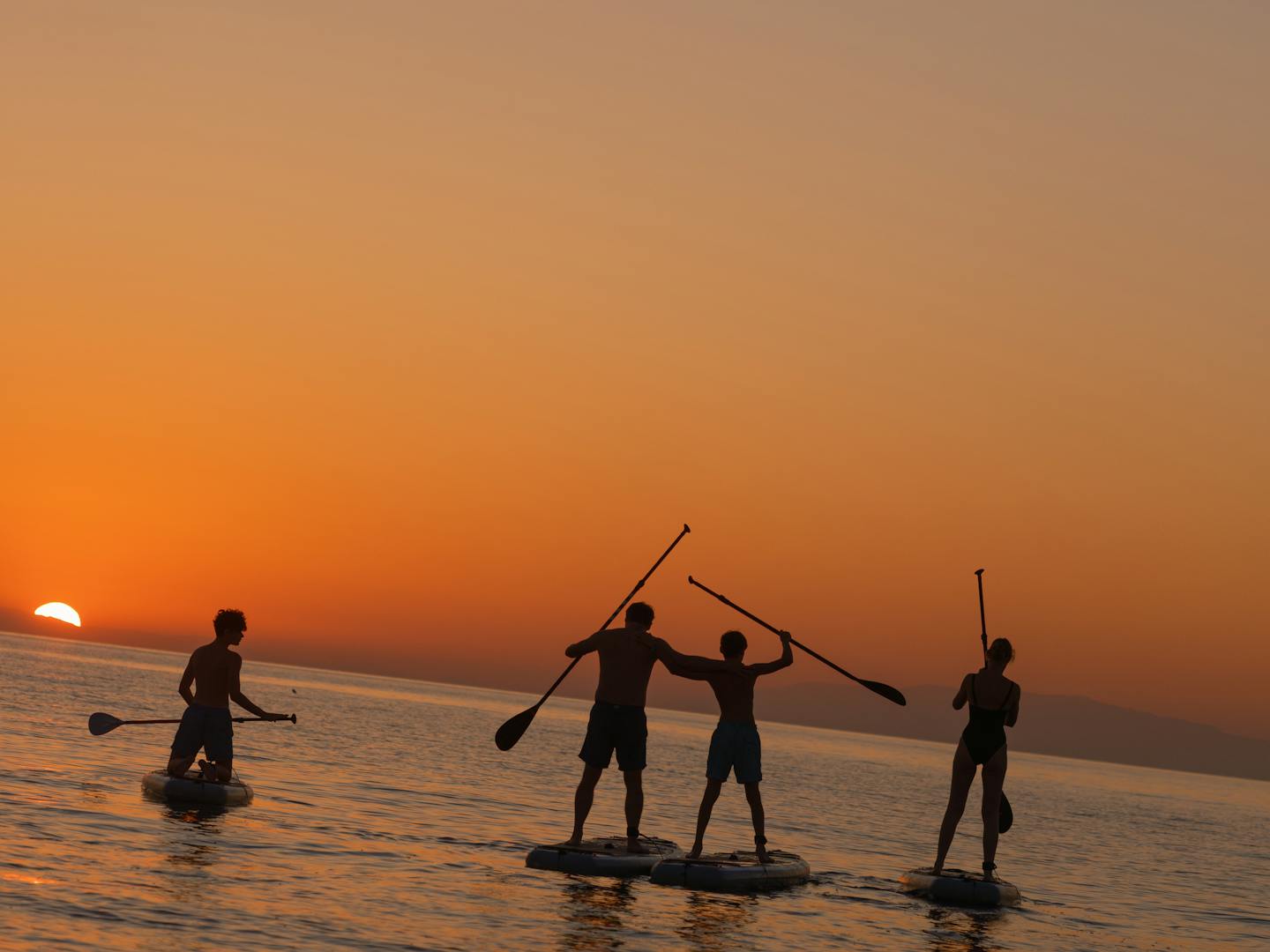Silhouettes of four people stand-up paddleboarding at sunset on calm water. Three people are on paddleboards while one person stands in the shallow water. They're holding their paddles in various positions against a vibrant orange and amber sky as the sun sets on the horizon, creating dramatic backlighting and reflections on the water's surface.