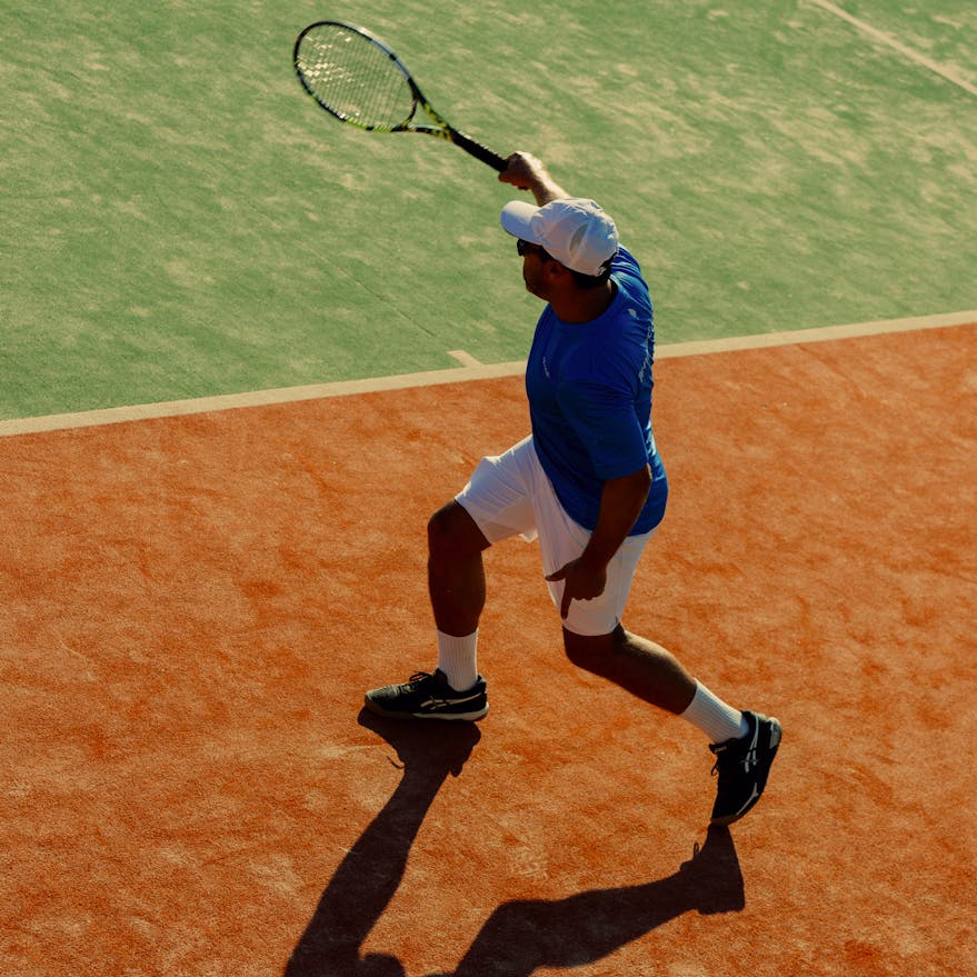 Overhead view of a tennis player mid-swing on a clay court, wearing a blue shirt, white shorts, white cap, and black shoes. The player holds a tennis racket raised above their head, casting a distinct shadow on the orange-red clay surface. The adjacent grass court is visible in the background, creating a contrast between the two playing surfaces.