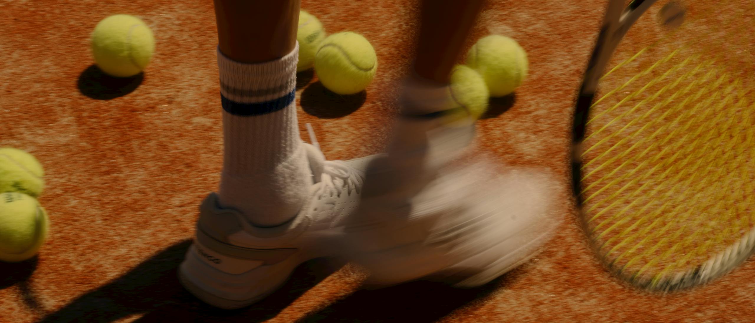 Motion-blurred action shot of a tennis player's legs and feet on a clay court, wearing white athletic shoes and striped socks with black shorts. Multiple tennis balls are scattered across the orange-red clay surface, and a tennis racket with yellow strings is visible on the right side. The player's shadow is cast prominently on the court, creating dramatic contrast.