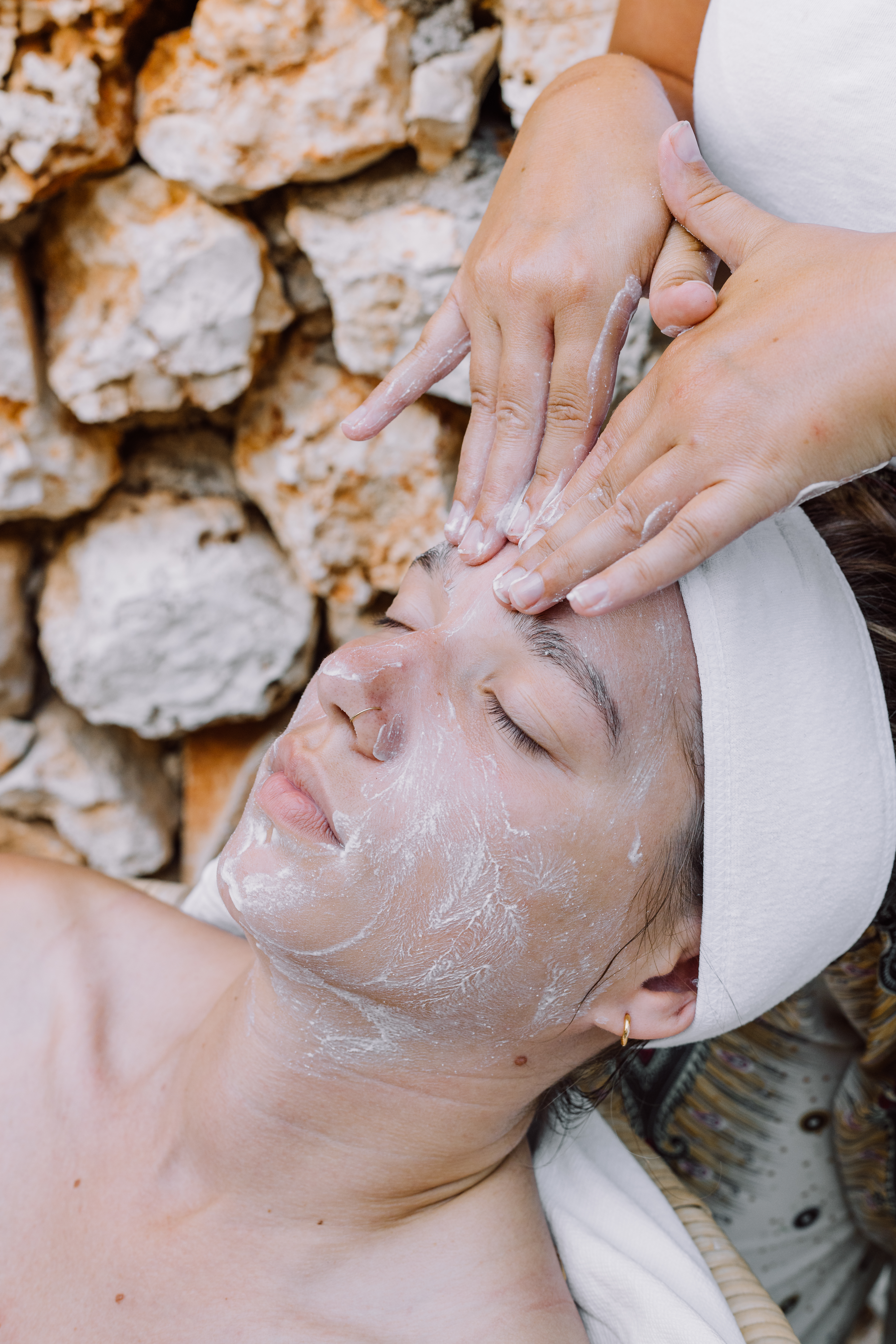 A woman receiving a facial treatment lies on a patterned mat against a textured stone wall background. An aesthetician's hands gently apply a clear treatment or serum to the client's face, which is covered with a translucent facial product. The client wears a white headband and has her eyes peacefully closed during the spa treatment.