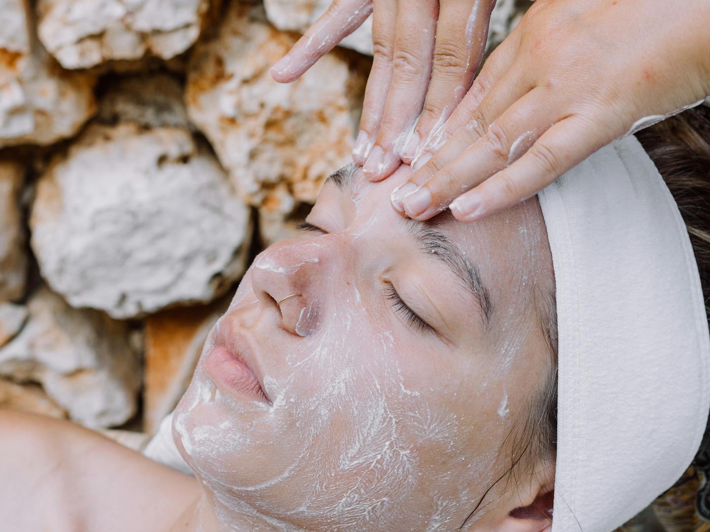 A woman receiving a facial treatment lies on a patterned mat against a textured stone wall background. An aesthetician's hands gently apply a clear treatment or serum to the client's face, which is covered with a translucent facial product. The client wears a white headband and has her eyes peacefully closed during the spa treatment.