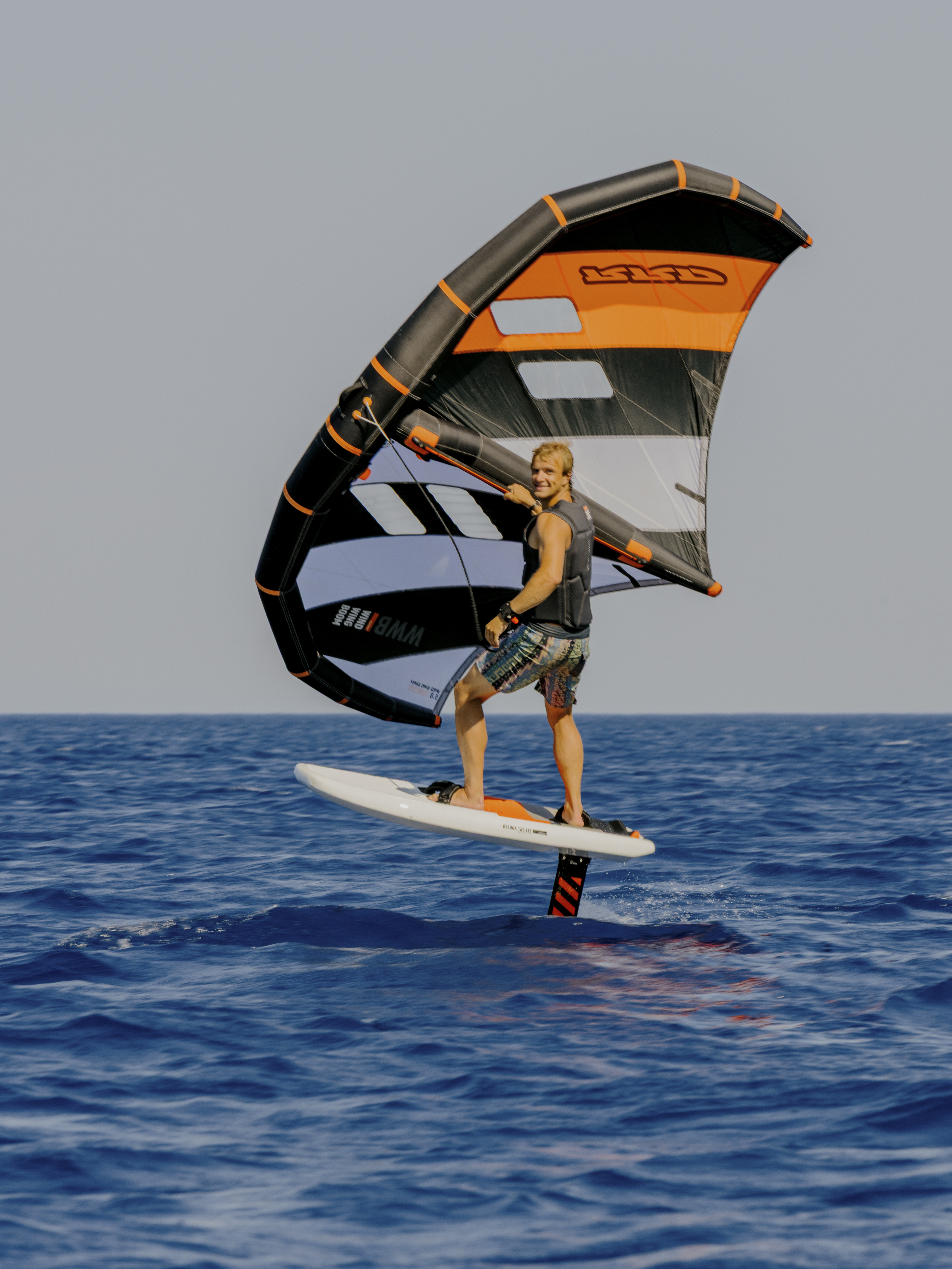 A wing foiler rides above the water on a hydrofoil board, holding a large wing sail with orange, gray, and black stripes. The rider wears a black vest and patterned shorts while gliding over calm, deep blue ocean water against a clear sky.