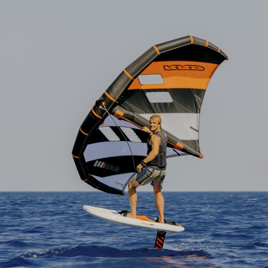 A wing foiler rides above the water on a hydrofoil board, holding a large wing sail with orange, gray, and black stripes. The rider wears a black vest and patterned shorts while gliding over calm, deep blue ocean water against a clear sky.