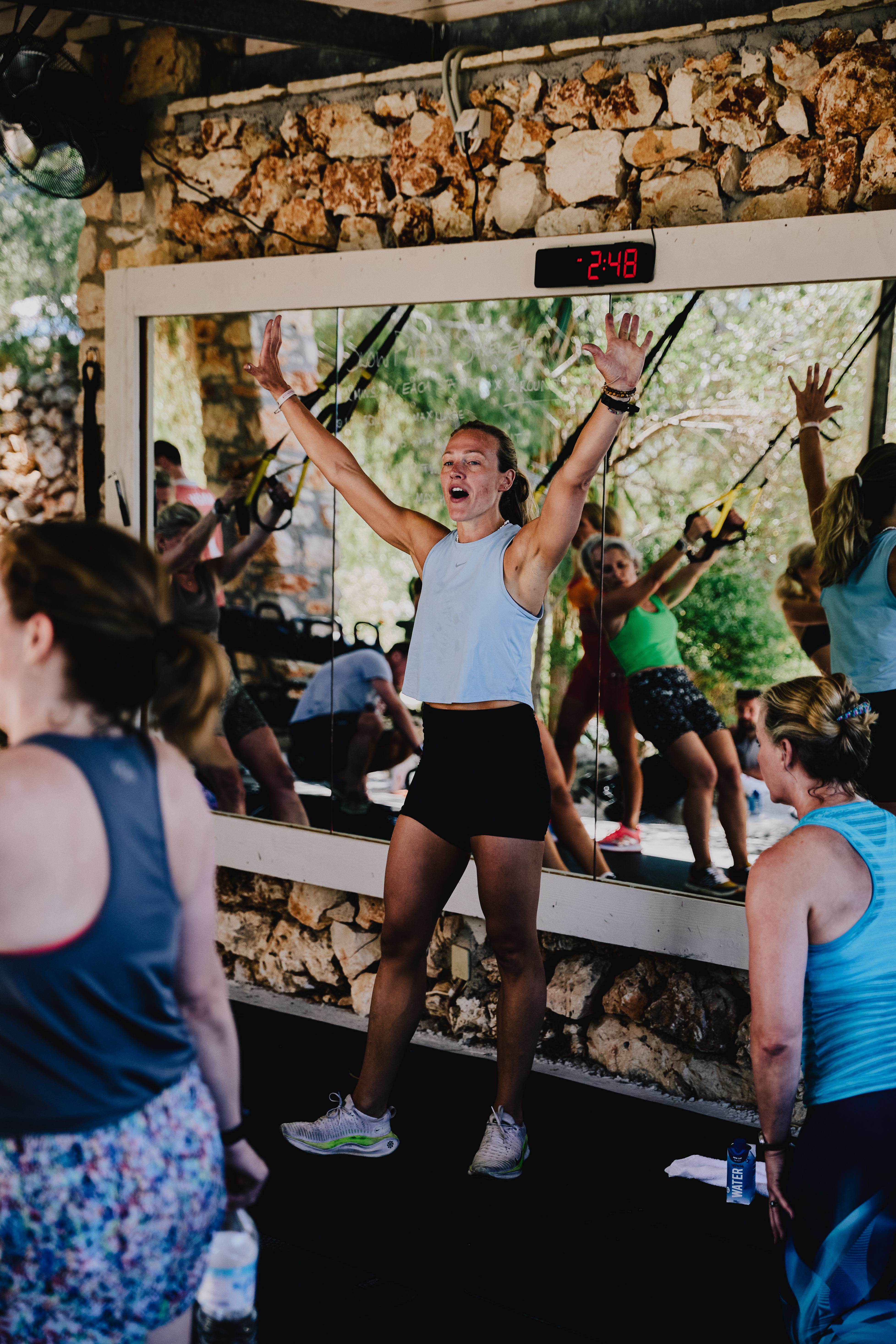 An energetic outdoor fitness class taking place under a rustic stone-walled shelter. A woman in a light blue tank top and black shorts leads the session with arms raised enthusiastically in front of a large mirror displaying "2:48" on a digital timer above. Participants in workout attire follow along, visible both directly and reflected in the mirror. The natural setting features exposed stone walls and views of greenery in the background.