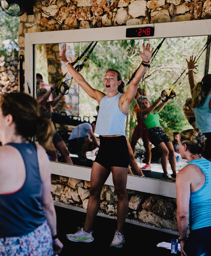 An energetic outdoor fitness class taking place under a rustic stone-walled shelter. A woman in a light blue tank top and black shorts leads the session with arms raised enthusiastically in front of a large mirror displaying "2:48" on a digital timer above. Participants in workout attire follow along, visible both directly and reflected in the mirror. The natural setting features exposed stone walls and views of greenery in the background.