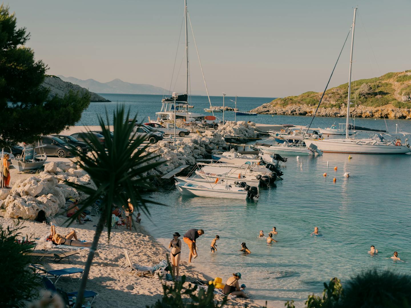 A scenic Mediterranean coastal cove during golden hour, featuring a small marina with sailboats and smaller vessels docked along a rocky breakwater. Beachgoers wade in the calm turquoise water while others relax on the sandy beach. Palm trees and lush vegetation frame the scene, with mountains visible across the tranquil sea in the distance under a clear sky.