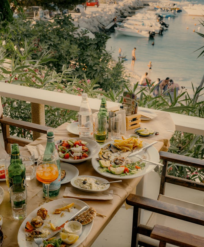 An outdoor waterfront dining table set with Mediterranean cuisine including Greek salad, grilled fish with lemon, french fries, and seafood appetizers. Green glass bottles and colorful drinks accompany the meal. Beyond the white railing with lush greenery, a marina with white boats is visible, and beachgoers can be seen in the background during golden hour lighting.