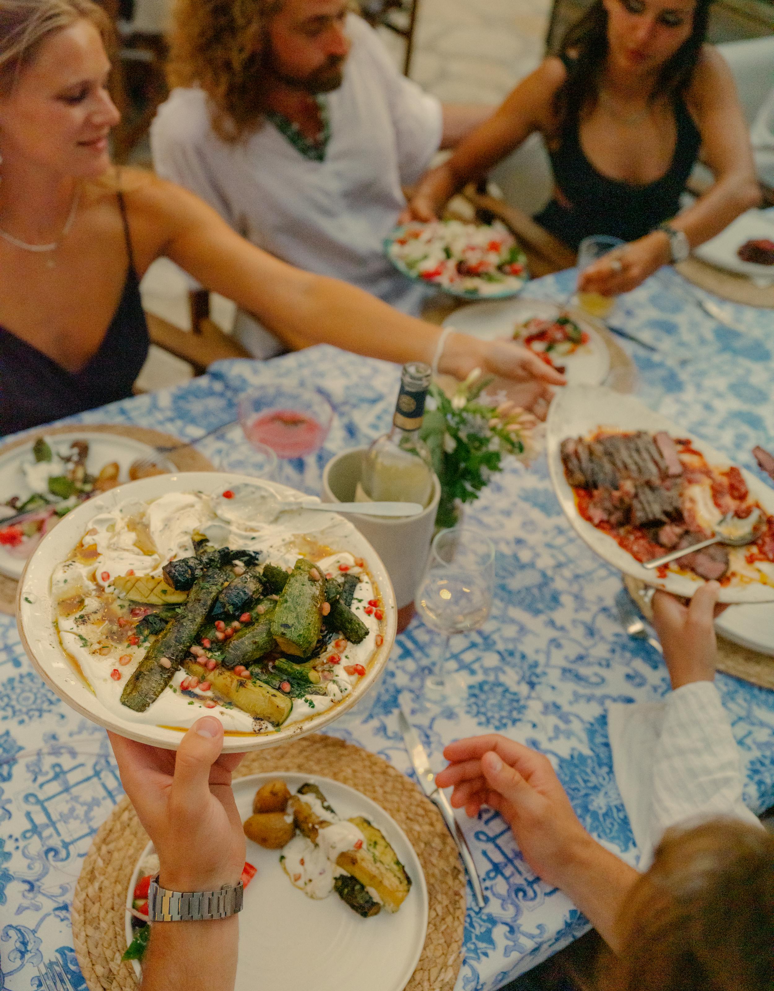A group of people gathered around a table with a blue and white patterned tablecloth, sharing a Mediterranean-style meal family-style. The central focus is on hands holding a white plate with grilled zucchini topped with yogurt sauce, herbs, and chili flakes. The table features multiple shared dishes including grilled vegetables, salads with tomatoes, sliced meat, and small bowls of sauces. Guests in casual summer attire reach across the table to serve themselves in a warm, communal dining atmosphere.