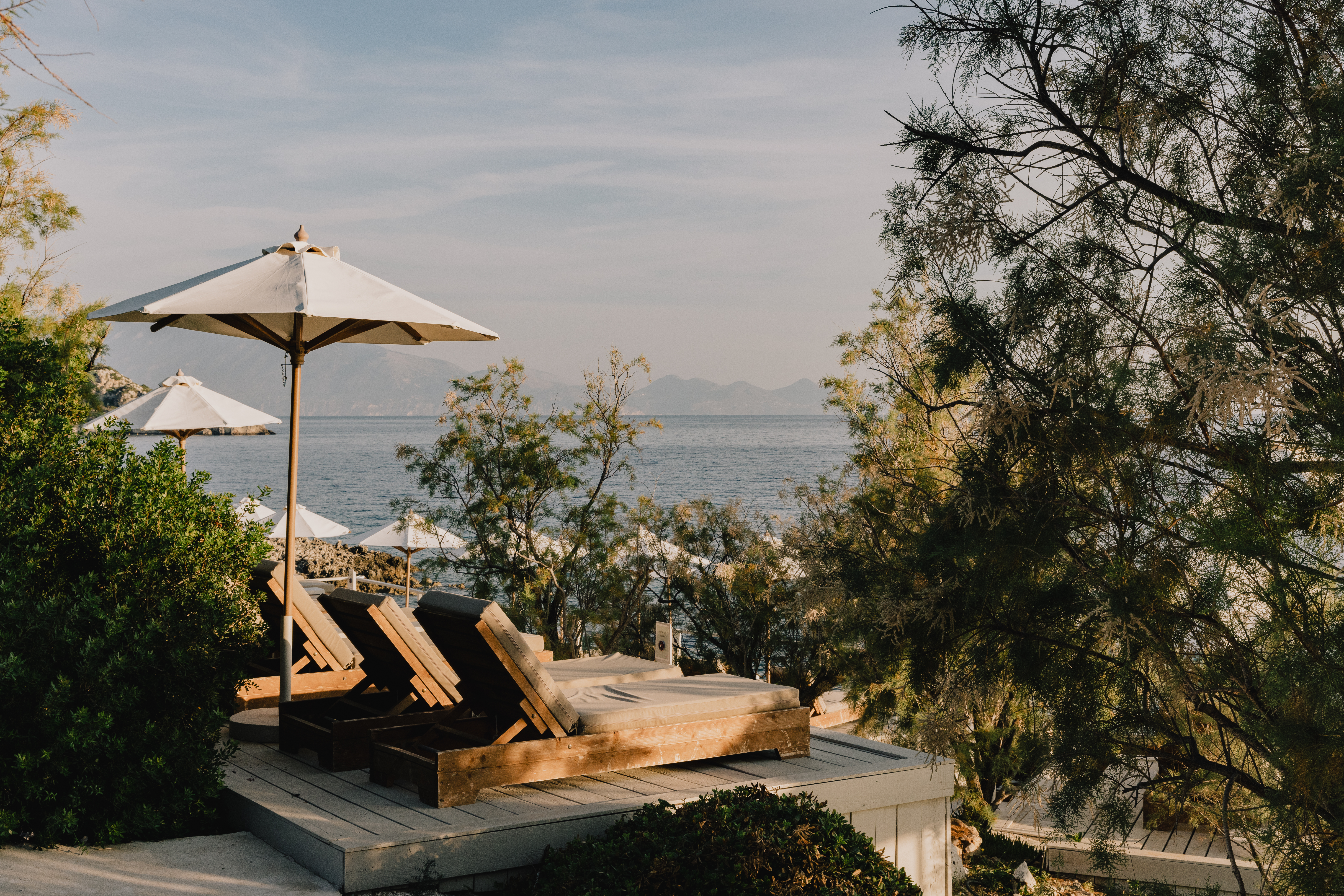 A serene coastal terrace featuring wooden sun loungers with white cushions and cream umbrellas overlooking a calm blue sea. The multi-level deck is surrounded by lush Mediterranean vegetation including pine trees and coastal shrubs. Mountains are visible across the water in the misty distance under a soft, overcast sky, creating a peaceful retreat atmosphere during golden hour lighting.