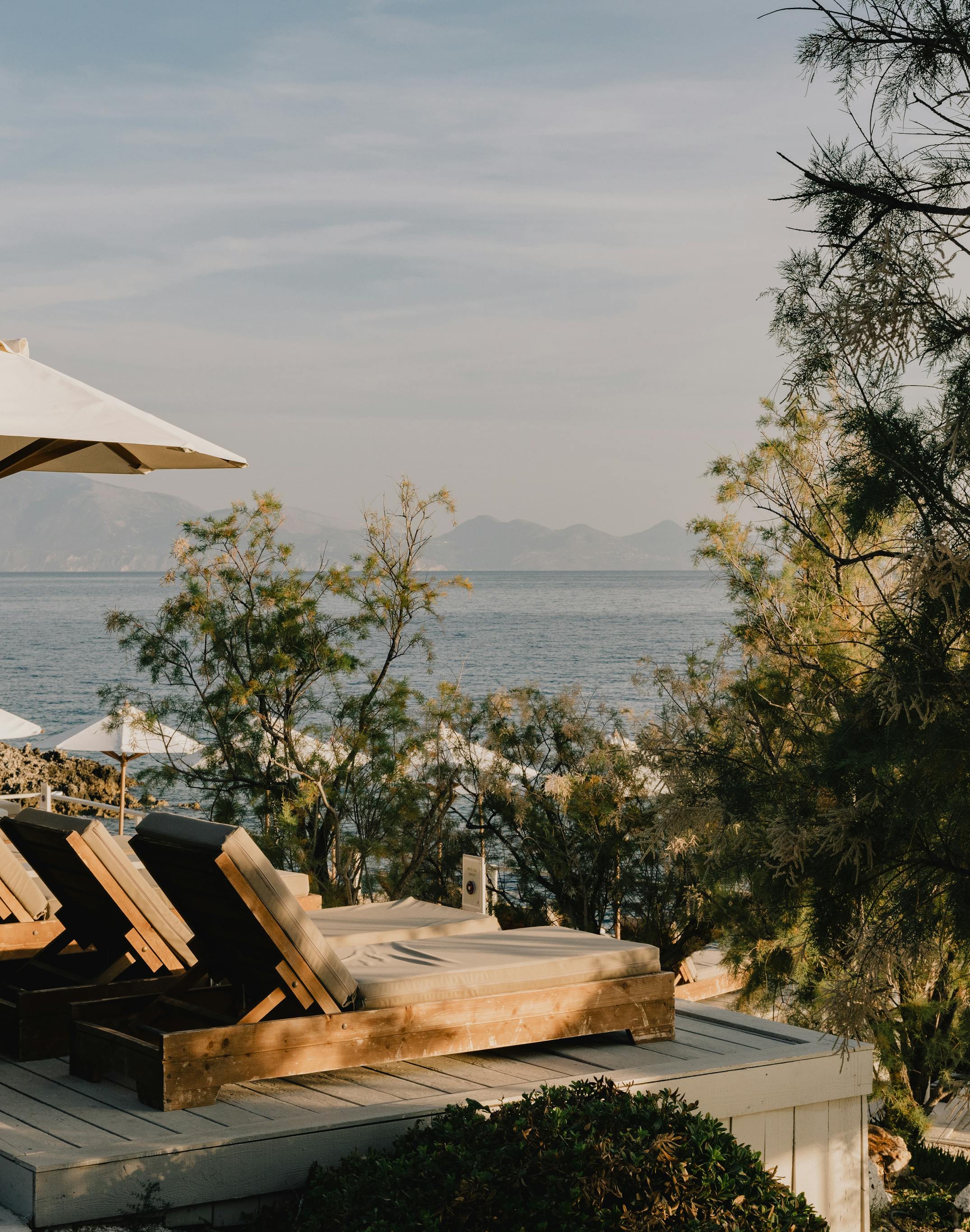 A serene coastal terrace featuring wooden sun loungers with white cushions and cream umbrellas overlooking a calm blue sea. The multi-level deck is surrounded by lush Mediterranean vegetation including pine trees and coastal shrubs. Mountains are visible across the water in the misty distance under a soft, overcast sky, creating a peaceful retreat atmosphere during golden hour lighting.