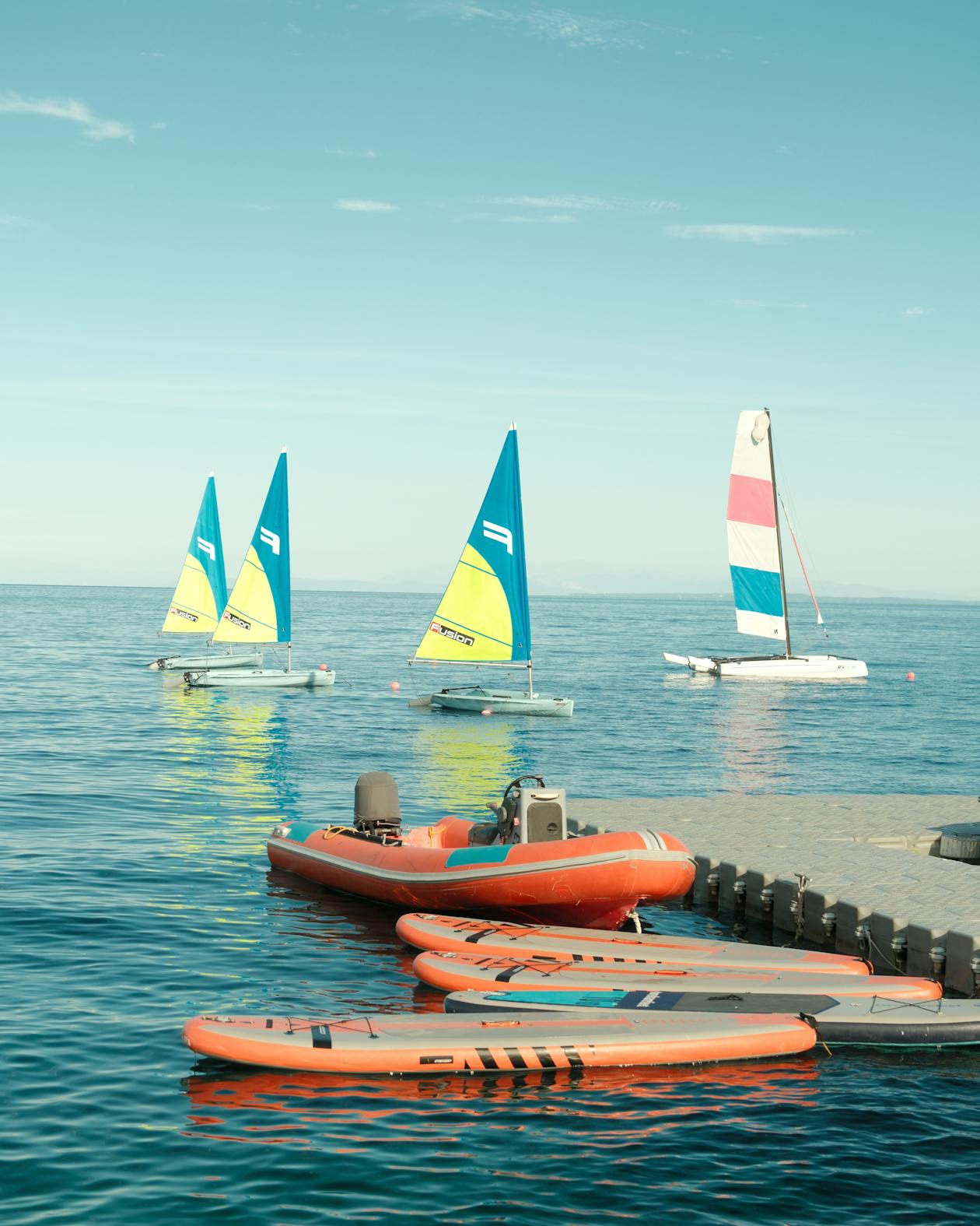 A serene water sports scene showing three small catamarans with colorful sails (turquoise with yellow accents, and white with pink/blue stripes) anchored in calm blue water. In the foreground, an orange inflatable boat and several orange paddleboards or hydrofoils are lined up at the water's edge. The clear sky and tranquil water create a perfect setting for water activities.