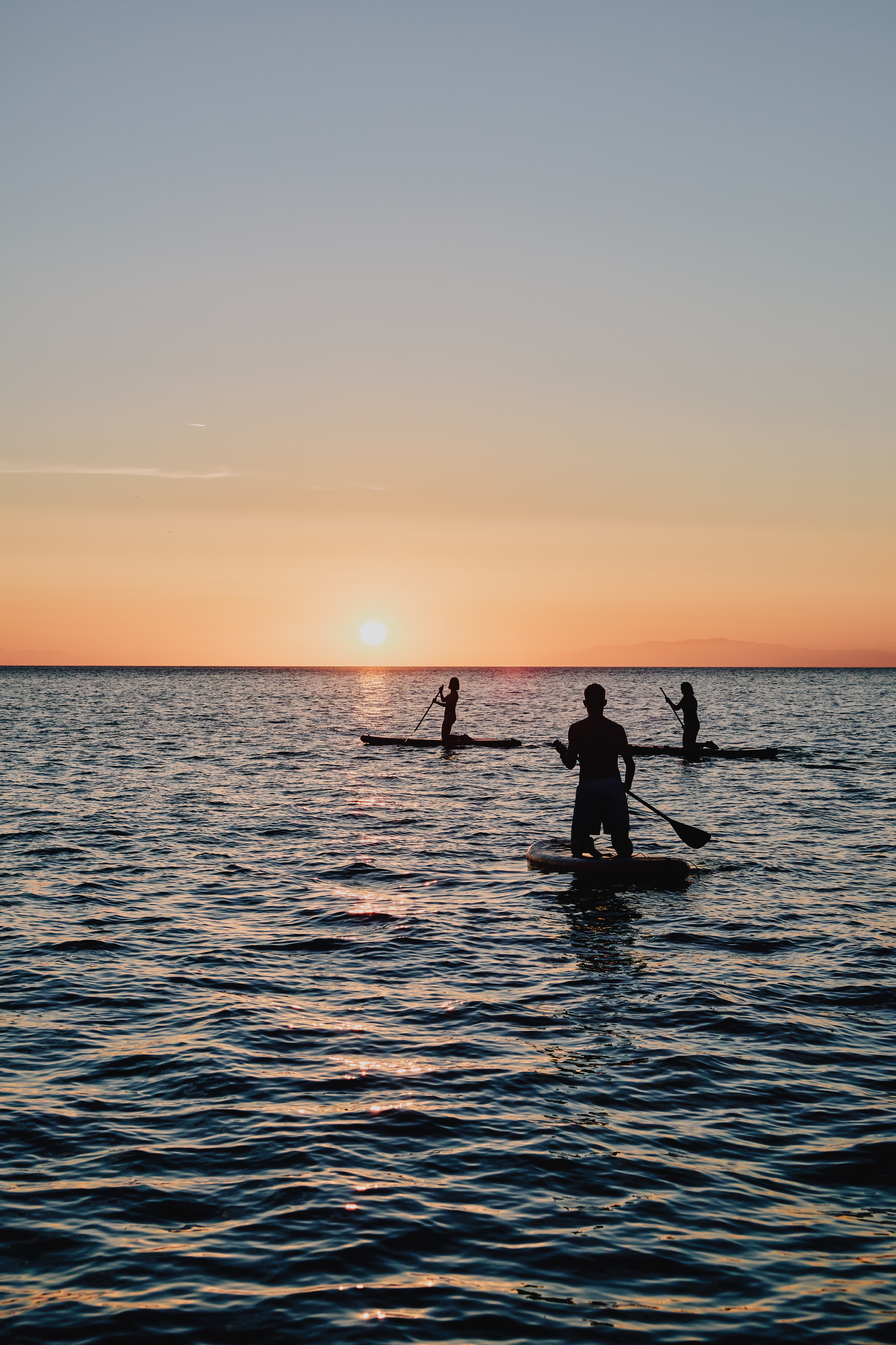Silhouettes of three stand-up paddleboarders on calm water during sunset or sunrise. The foreground shows one paddler in sharp silhouette, while two others are visible in the distance. The sky features a gradient from peachy orange at the horizon to soft blue-gray above, with the sun visible as a bright orb near the horizon. The water surface reflects the warm colors of the sky, creating gentle ripples throughout the scene.