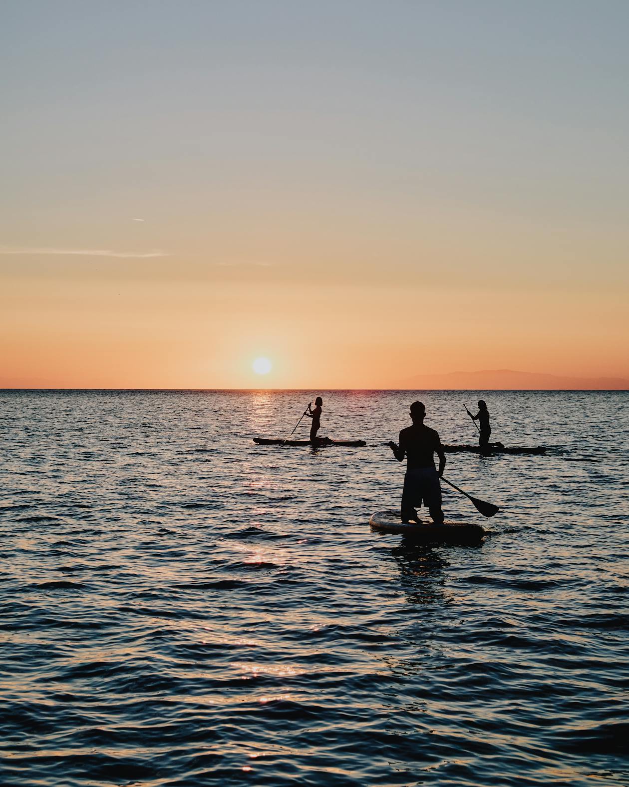 Silhouettes of three stand-up paddleboarders on calm water during sunset or sunrise. The foreground shows one paddler in sharp silhouette, while two others are visible in the distance. The sky features a gradient from peachy orange at the horizon to soft blue-gray above, with the sun visible as a bright orb near the horizon. The water surface reflects the warm colors of the sky, creating gentle ripples throughout the scene.