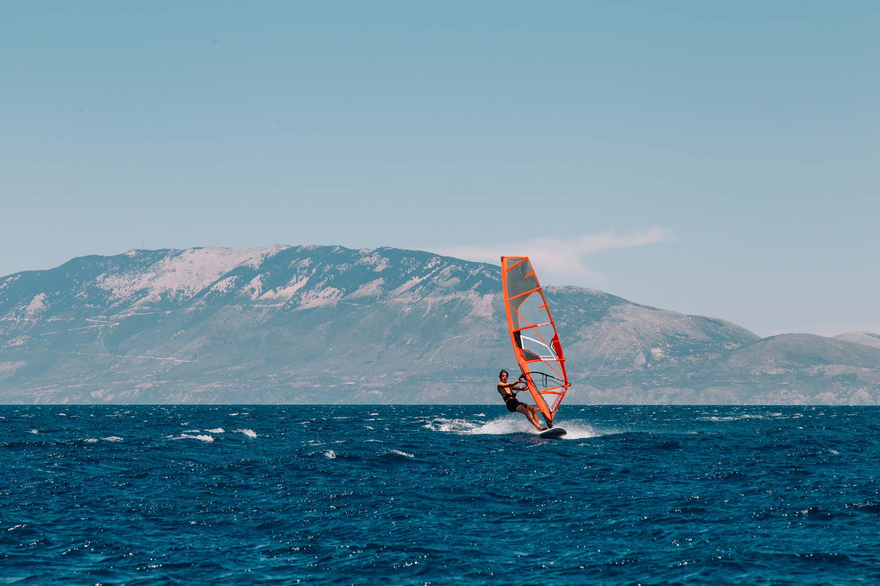 A windsurfer riding choppy blue waters with a bright orange and white sail, set against a dramatic backdrop of mountains with snow-dusted peaks. White spray kicks up around the board as the surfer navigates the waves under a pale blue sky with wispy clouds.