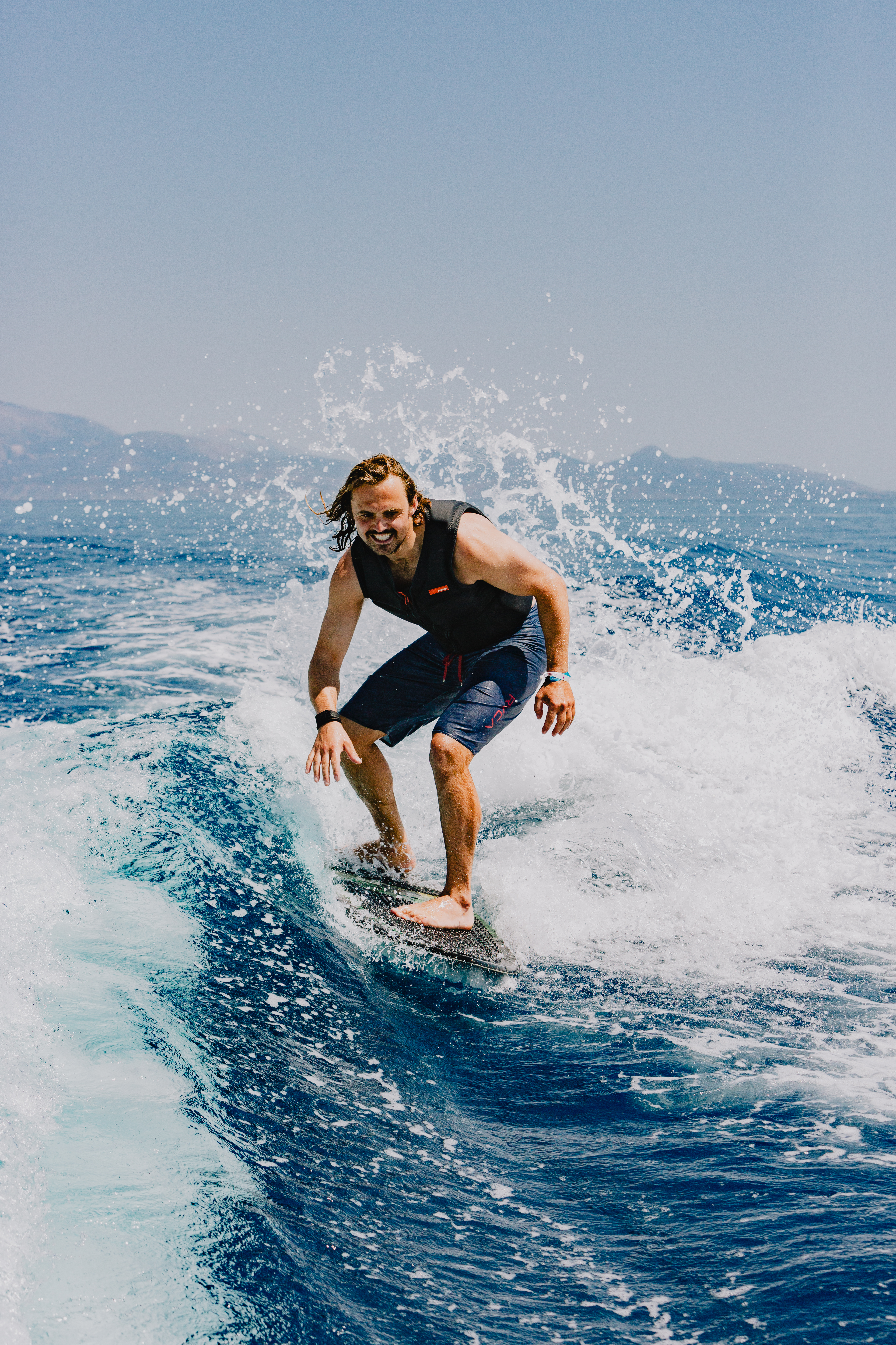 A smiling surfer riding a wave on a blue surfboard, wearing a black wetsuit vest and blue board shorts. Water sprays dramatically around them as they navigate the white foam on the deep blue ocean water. Mountains are visible in the hazy background under a clear sky.