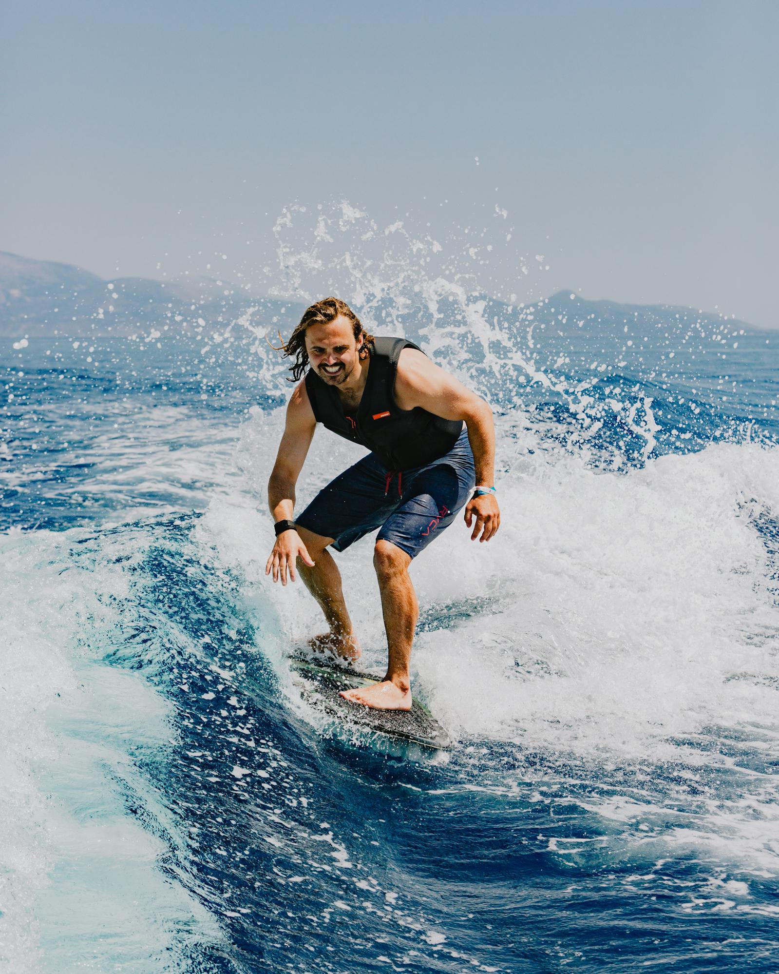 A smiling surfer riding a wave on a blue surfboard, wearing a black wetsuit vest and blue board shorts. Water sprays dramatically around them as they navigate the white foam on the deep blue ocean water. Mountains are visible in the hazy background under a clear sky.