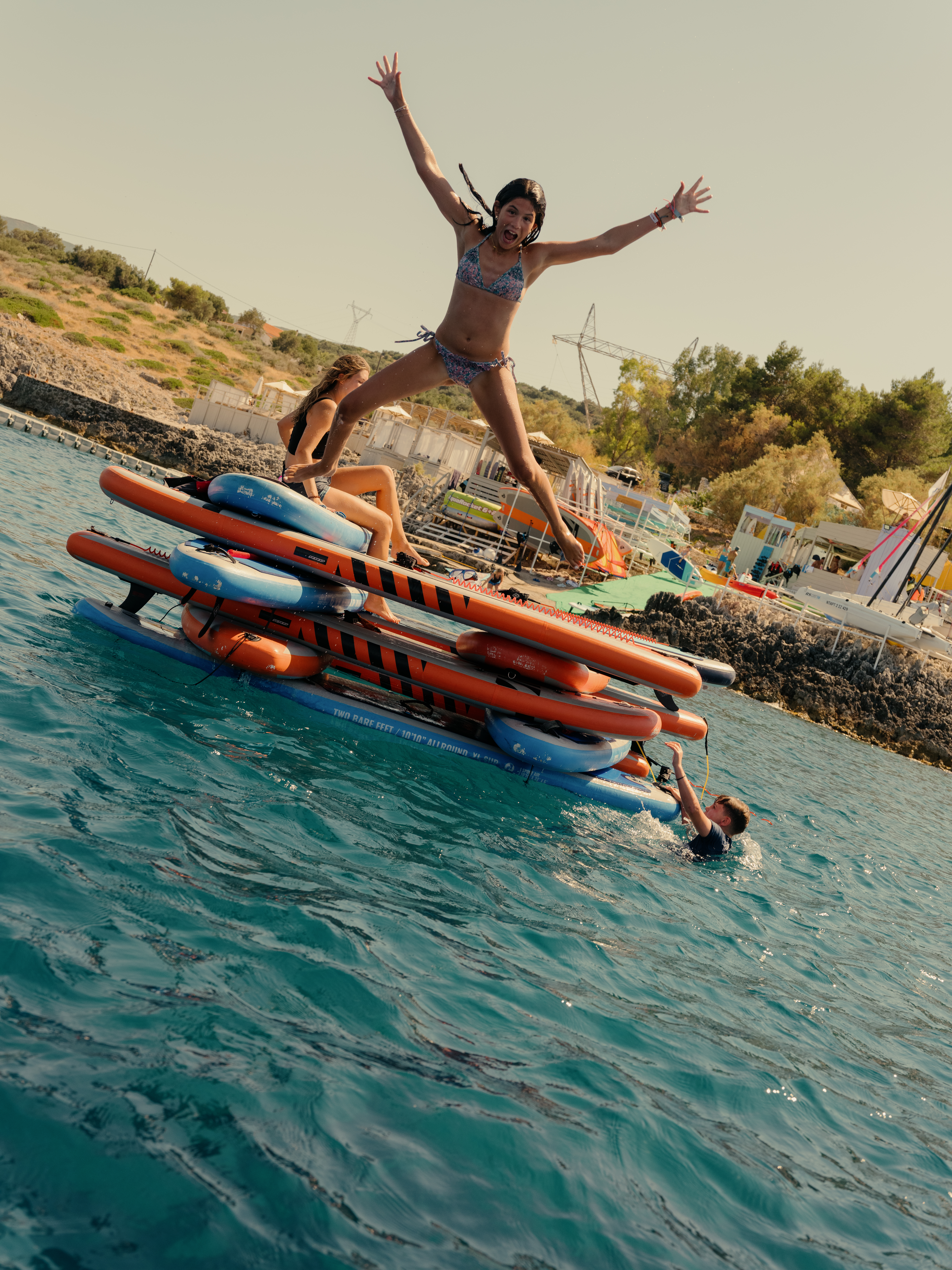 A girl in a patterned bikini jumps joyfully with arms spread wide from a floating inflatable obstacle course, while others lounge on the structure in the turquoise water. The coastal setting shows a hillside beach area with buildings, greenery, and equipment in the background. Someone swims in the water below the jump platform, which features orange and blue striped bars.