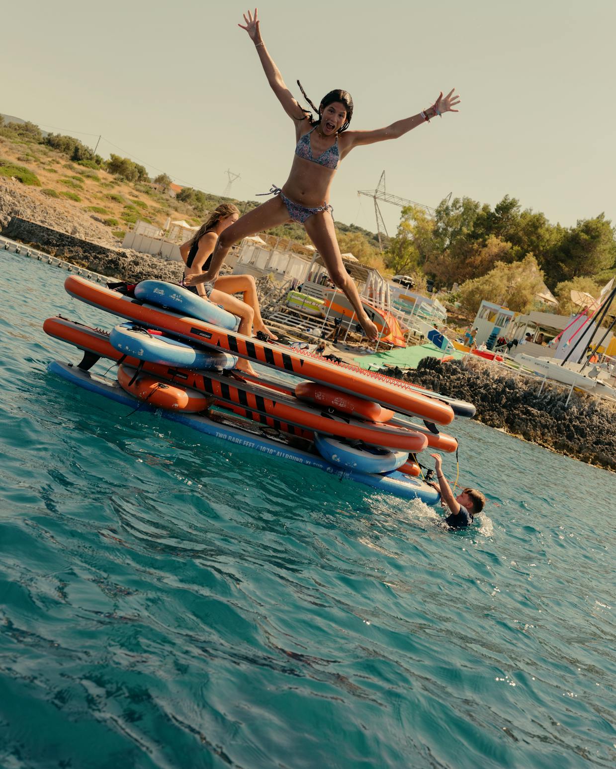 A girl in a patterned bikini jumps joyfully with arms spread wide from a floating inflatable obstacle course, while others lounge on the structure in the turquoise water. The coastal setting shows a hillside beach area with buildings, greenery, and equipment in the background. Someone swims in the water below the jump platform, which features orange and blue striped bars.