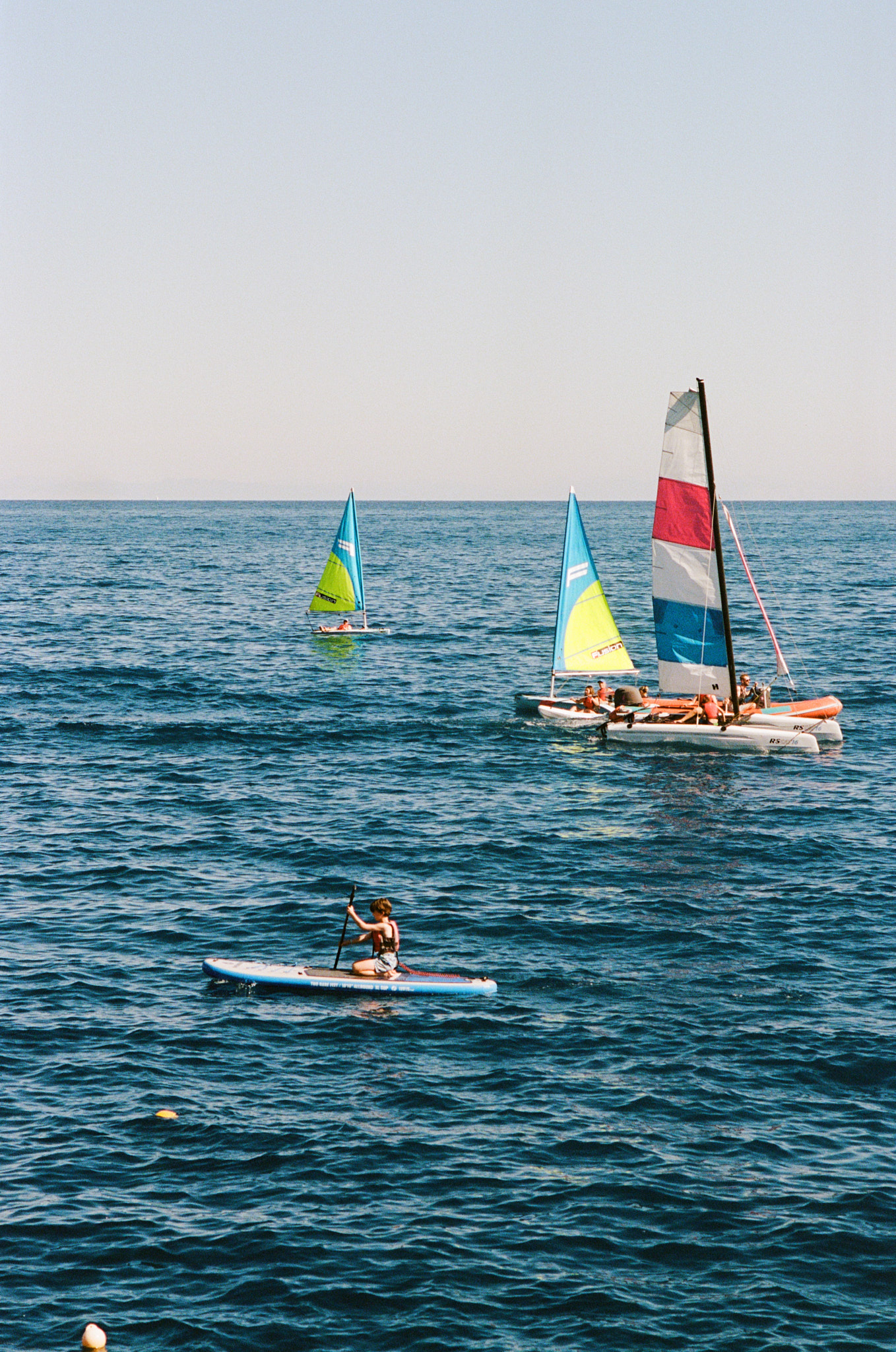 A serene water sports scene on calm blue water featuring multiple watercraft - a paddleboarder on a blue SUP board in the foreground, a small windsurfer with a yellow and blue sail in the middle distance, and a larger orange catamaran with a red, white, and blue sail in the background. The horizon shows distant mountains under a hazy sky, with scattered yellow buoys visible in the water.