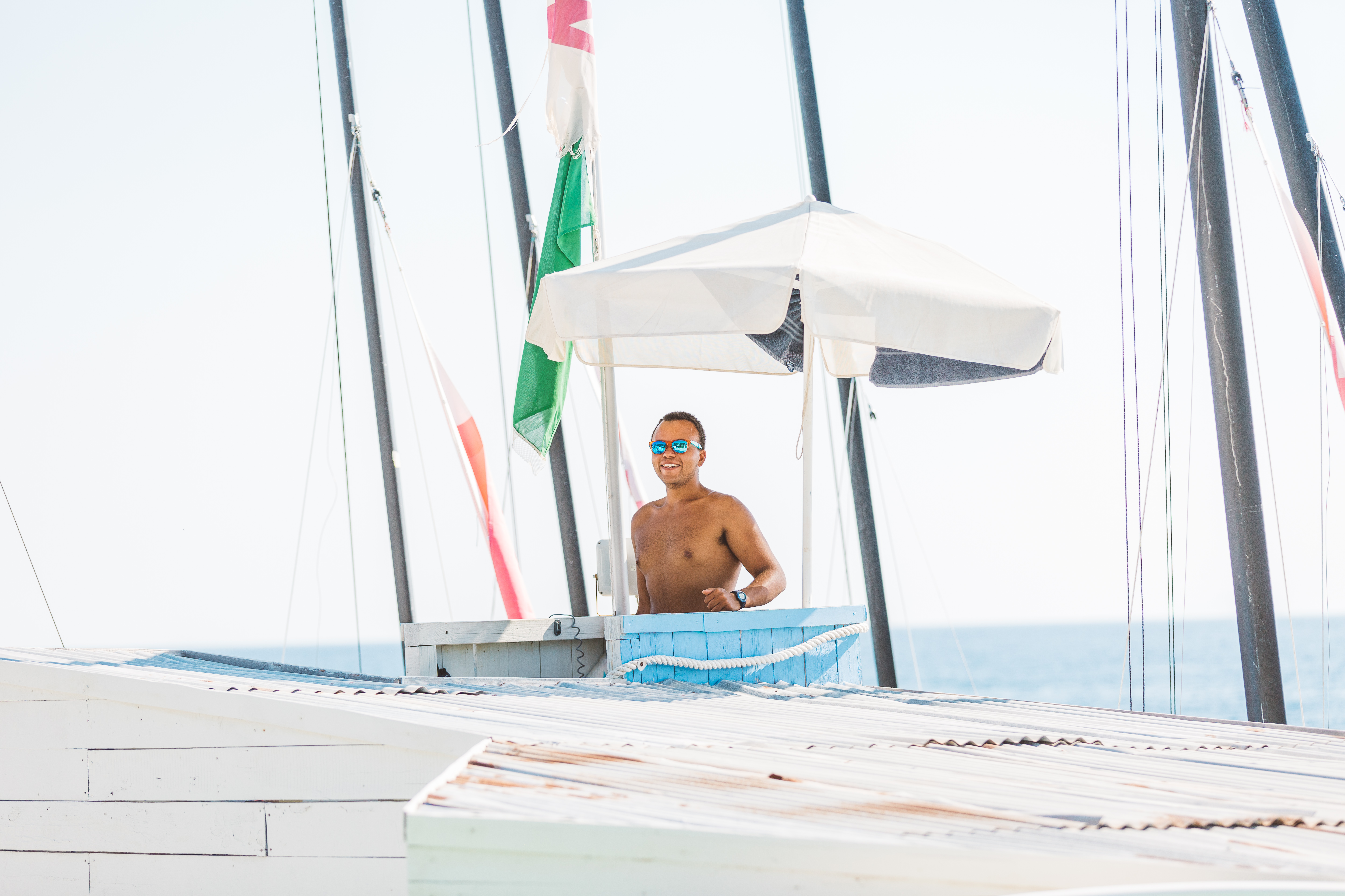 A shirtless man wearing blue mirrored sunglasses smiles from inside a white beach kiosk or towel station with an open-air design. The structure has a white canopy roof, blue towels visible on the counter, and sailing masts with rigging visible in the background against a bright, overcast sky. The scene appears to be at a waterfront or marina location.