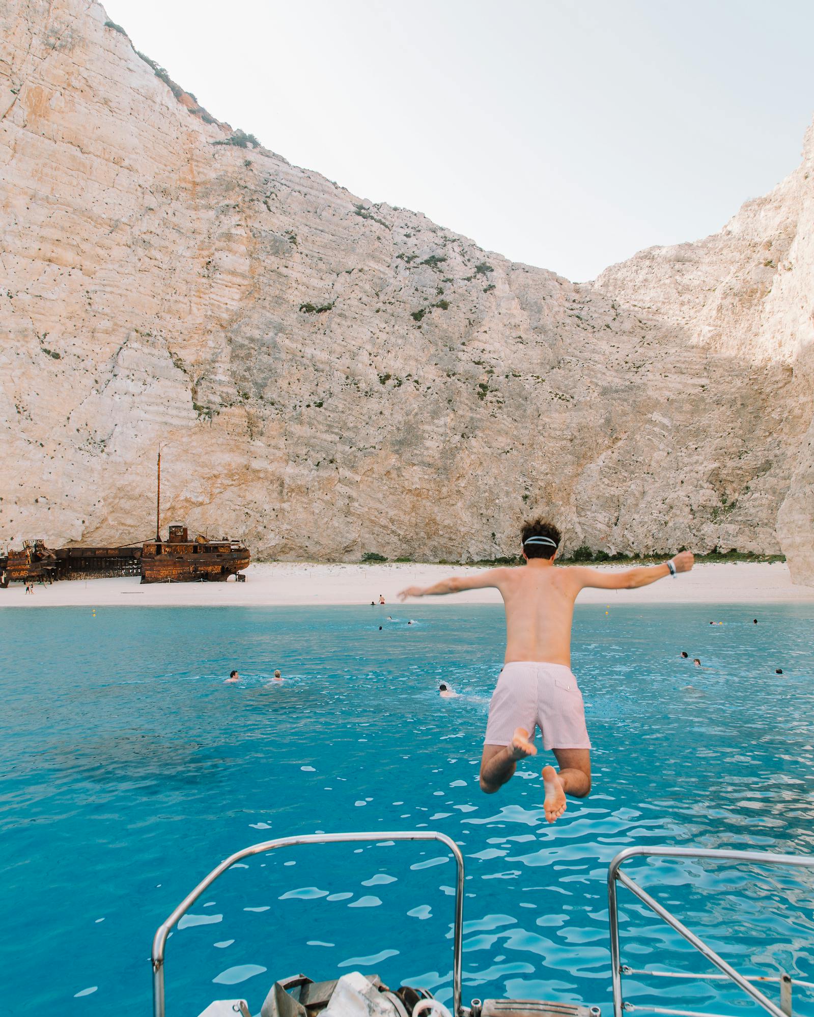A person mid-jump diving into bright turquoise Mediterranean waters from a boat's stern platform. The dramatic Navagio Beach (Shipwreck Beach) in Zakynthos, Greece serves as the backdrop, with its iconic towering white limestone cliffs framing the secluded cove. A traditional shipwreck is visible on the sandy beach, and the boat's metal railing is visible in the foreground.