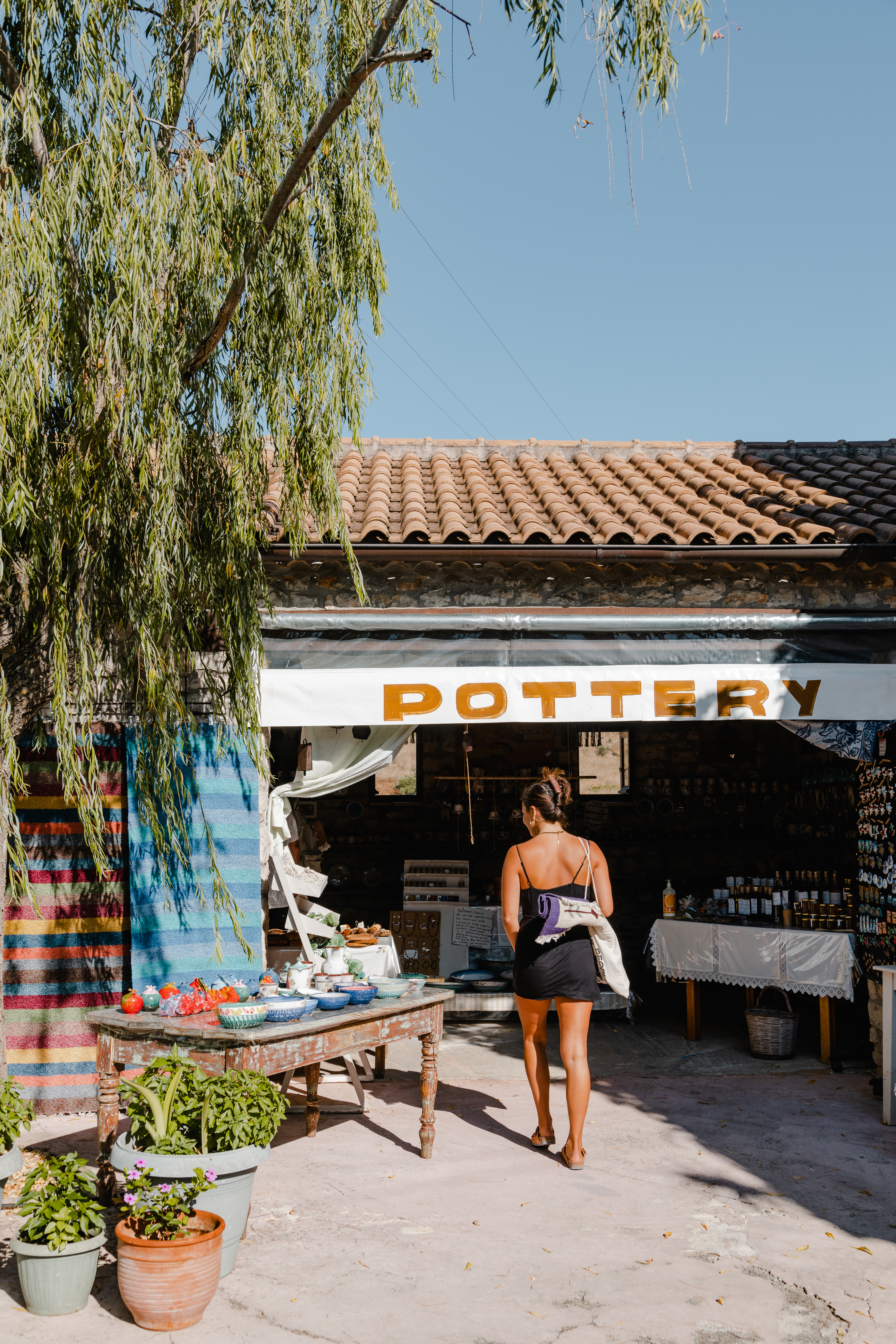 A woman in a black sundress walks toward a rustic pottery shop with a terracotta tile roof and white walls. The shop has 'POTTERY' signage in yellow letters above the entrance, where various ceramic items are displayed. A large tree with drooping branches provides natural shade over the concrete courtyard, which features several potted plants. The Mediterranean-style setting is captured in natural daylight.