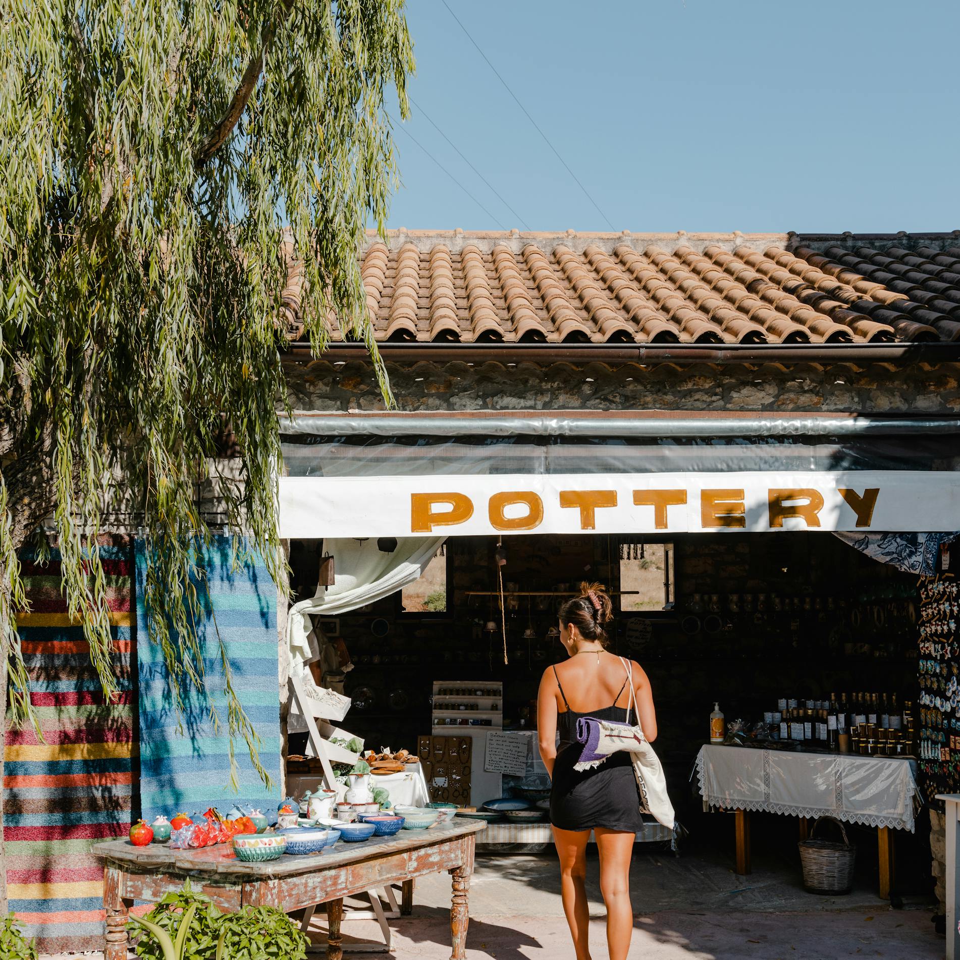 A woman in a black sundress walks toward a rustic pottery shop with a terracotta tile roof and white walls. The shop has 'POTTERY' signage in yellow letters above the entrance, where various ceramic items are displayed. A large tree with drooping branches provides natural shade over the concrete courtyard, which features several potted plants. The Mediterranean-style setting is captured in natural daylight.