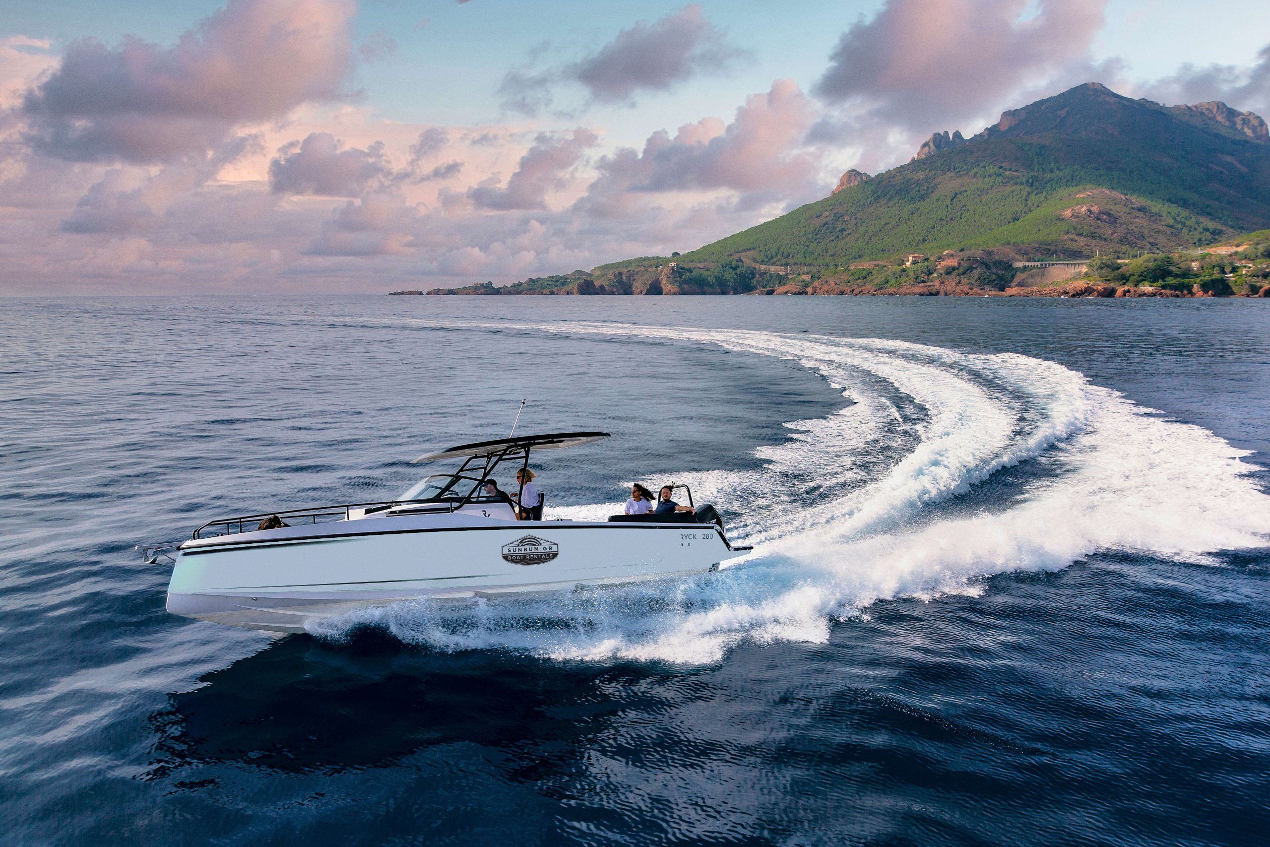 A white luxury powerboat speeds through calm waters at dusk, creating dramatic white wake trails. The boat has a black T-top canopy and carries several passengers. In the background, a dramatic mountainous coastline covered in green vegetation rises against a sky filled with pink and purple clouds. The scene captures the golden hour ambiance of coastal boating near a scenic Mediterranean or tropical setting.