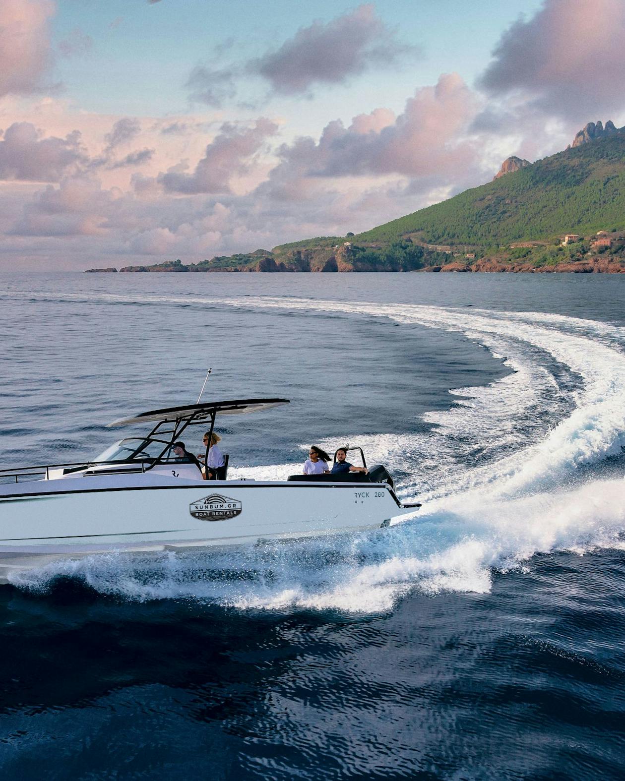 A white luxury powerboat speeds through calm waters at dusk, creating dramatic white wake trails. The boat has a black T-top canopy and carries several passengers. In the background, a dramatic mountainous coastline covered in green vegetation rises against a sky filled with pink and purple clouds. The scene captures the golden hour ambiance of coastal boating near a scenic Mediterranean or tropical setting.