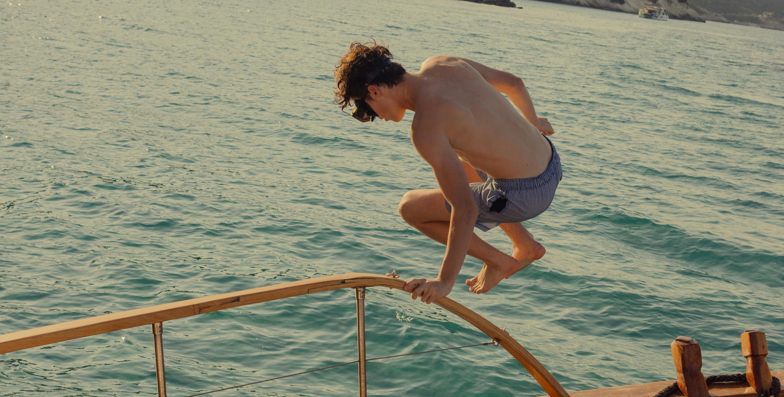A young person mid-jump from a wooden boat deck into turquoise coastal waters, with white cliffs and vegetation visible in the background. Another person with long wet hair watches from the boat's seating area. The boat features a classic wooden deck with teak flooring, white cushioned seating, metal railings, and nautical equipment. The scene captures a summer day of coastal leisure and water activities.