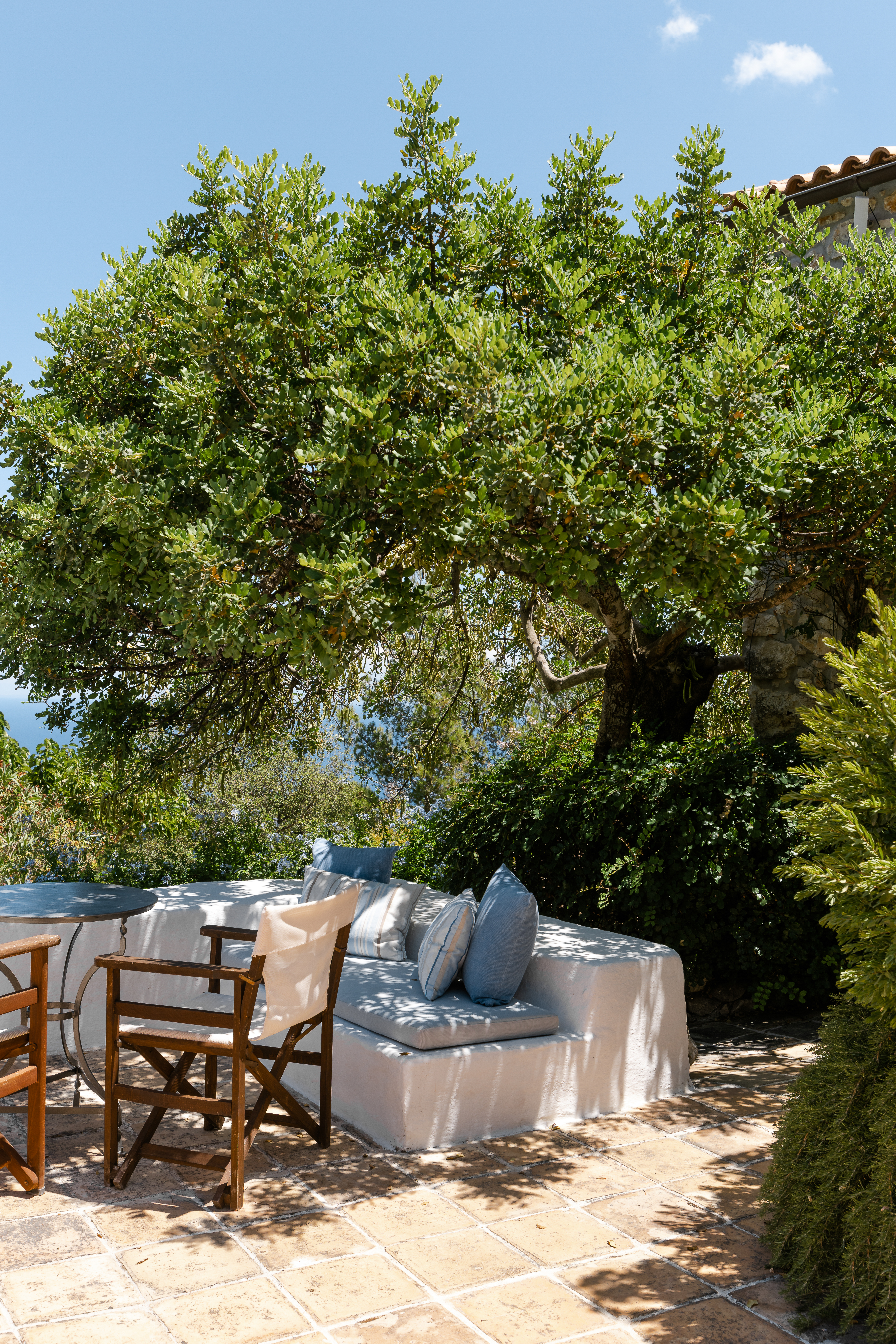 Shaded terrace seating with white cushioned sofa and wooden chairs beneath mature tree canopy. Dappled sunlight creates peaceful garden retreat with glimpse of red-tiled roof.