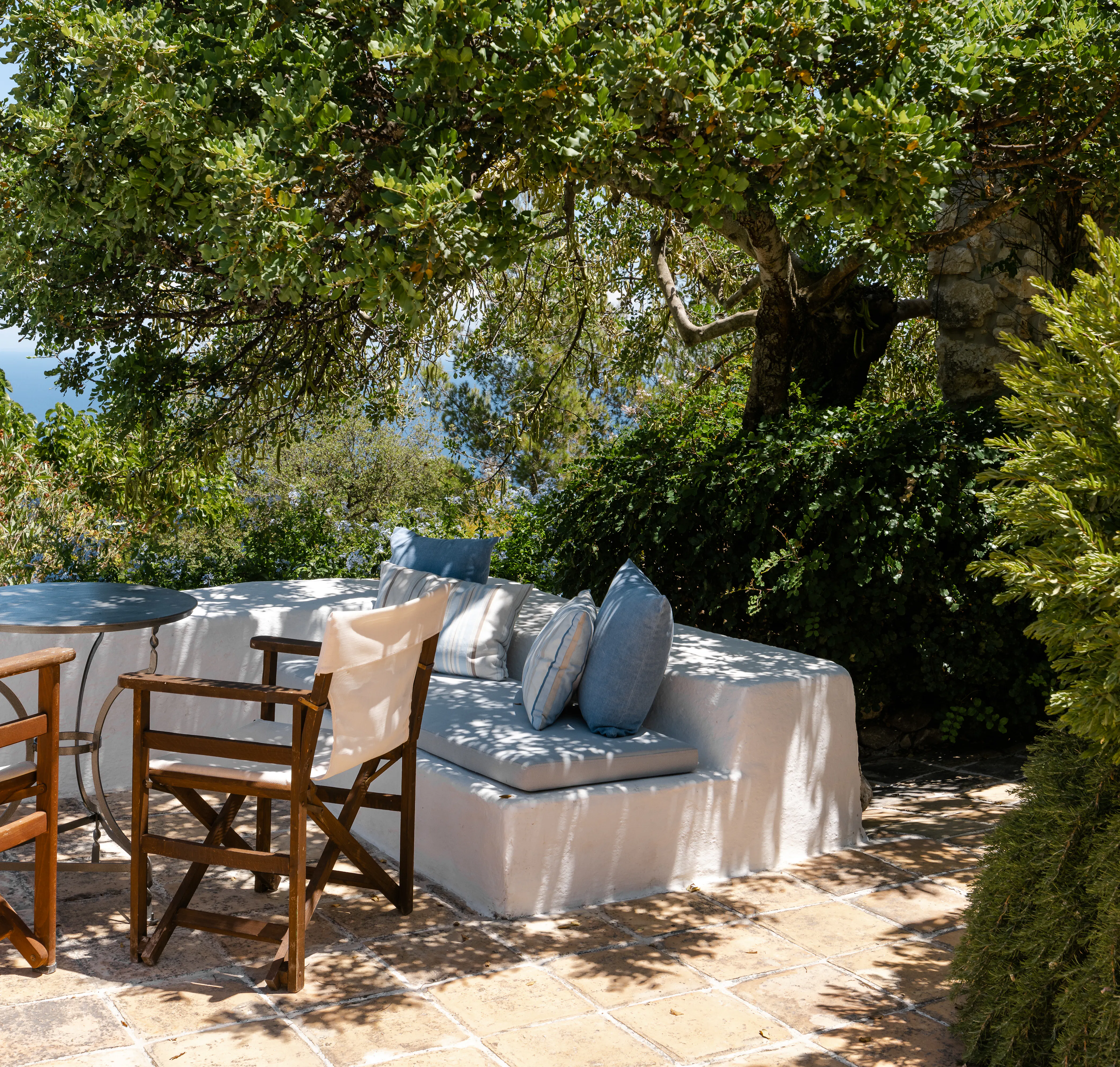 Shaded terrace seating with white cushioned sofa and wooden chairs beneath mature tree canopy. Dappled sunlight creates peaceful garden retreat with glimpse of red-tiled roof.